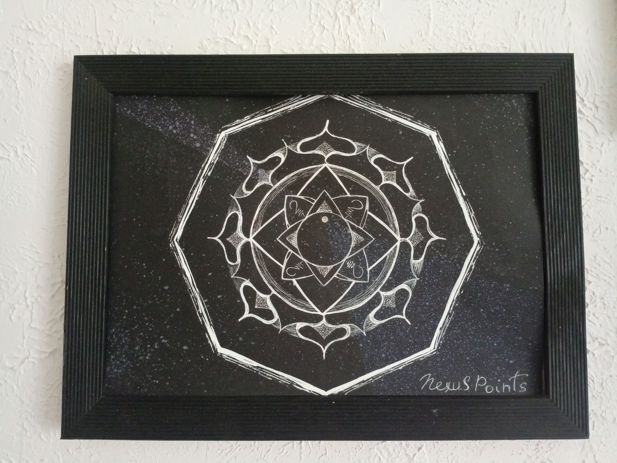 A framed black and white drawing of a mandala hangs on a textured white wall. The mandala is intricate with geometric shapes and decorative elements. The frame is black and ridged. In the bottom right corner of the drawing, the artist's signature "Nexus Points" is visible in white script. The image does not provide any information about its location in Saratov, Russia.