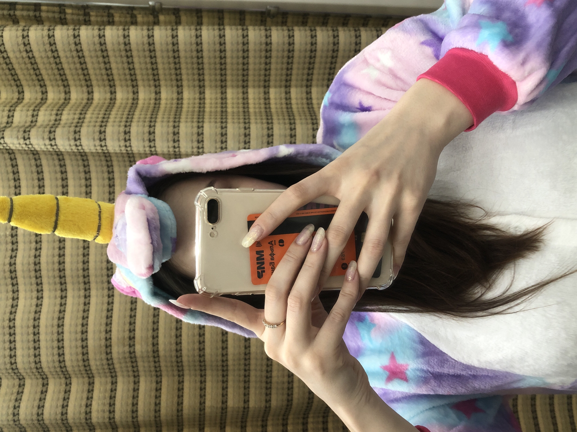 A person wearing a unicorn onesie is holding a cell phone in front of their face. A bank card is behind the cell phone. The person is on a striped, patterned couch. The location is not discernible in this image, and there is no way to identify the location as Chaikovsky, Russia.