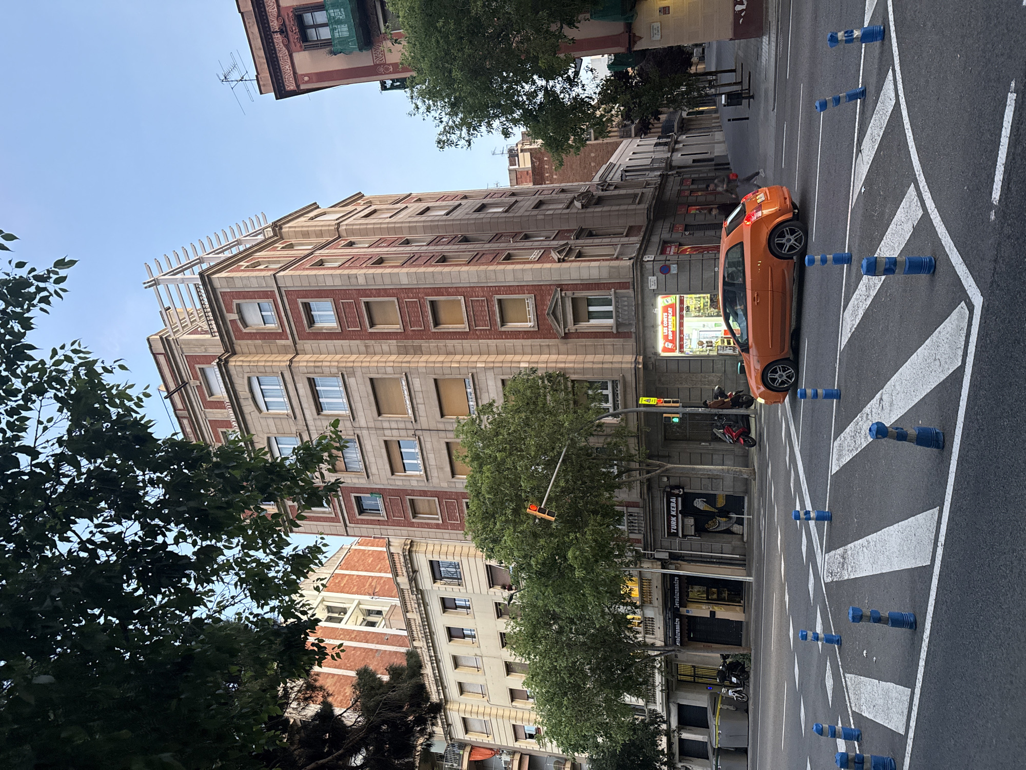 A brightly lit urban street scene in Barcelona, Spain, unfolds under a clear blue sky. The image captures a street lined with multi-story buildings, trees, and road infrastructure, observed during the daytime.
In the foreground and middle ground, an asphalt road displays distinct white chevron road markings and a zebra crosswalk. Several blue and white plastic traffic bollards are positioned along these markings. An orange Audi A3 hatchback is prominently parked on the street, near the crosswalk, while at least two motorbikes or scooters are visible parked further down.
The left side of the frame is dominated by a tall, multi-story building featuring a red and beige brick and stone facade, numerous windows with light-colored shutters, and decorative elements including a distinct finial-topped structure along its roofline. Another building with a lighter, possibly off-white or beige facade, stands adjacent to it. Lush green trees are interspersed along the street, with their foliage partially obscuring parts of the buildings and sky.
Visible commercial signage includes "LES CORTS SUPERMERCAT," "TURK KEBAI," and a smaller sign displaying "lavaropamandre.es." A traffic light showing a green signal hangs above the street. The scene depicts a typical, calm urban environment with clear weather conditions.