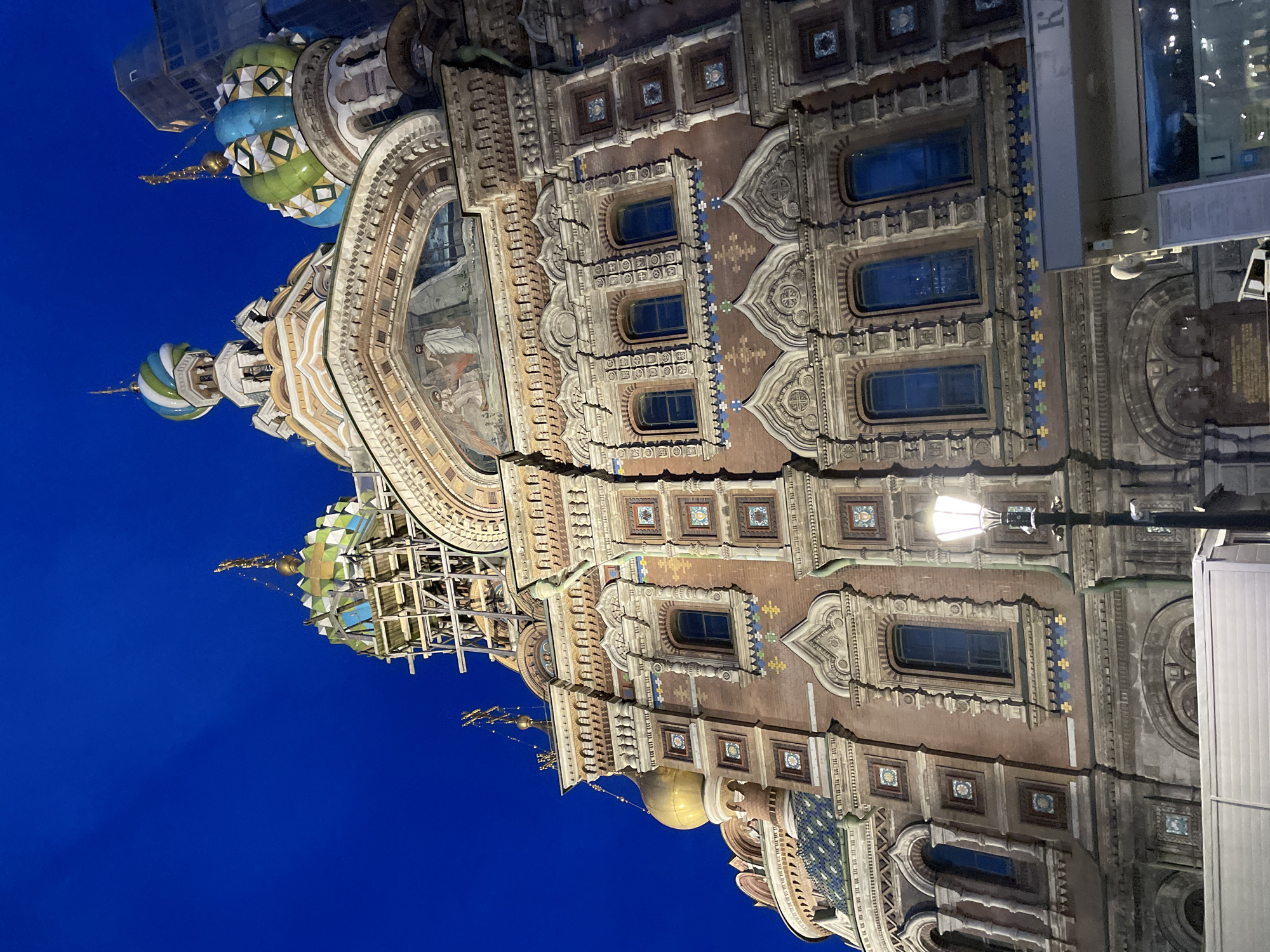 An elaborately decorated, multi-domed cathedral or church, characteristic of Russian Revival architecture, is depicted at night or deep twilight against a clear, dark blue sky. The building, likely the Church of the Savior on Spilled Blood in St. Petersburg, Russia, is richly adorned with intricate mosaic tiles, ornate brickwork, and stone carvings.
The facade features numerous arched windows, some with blue-tinted glass, and decorative panels in vibrant blue, green, and yellow patterns. A large arched portal showcases a detailed fresco or mosaic depicting figures. Several onion-shaped domes or cupolas top the structure, exhibiting colorful patterns in green, blue, white, and gold. One larger dome appears to be partially covered by scaffolding, suggesting maintenance or ongoing work. A smaller, glistening golden dome is also visible lower on the building.
The structure is brightly illuminated by artificial lights, highlighting its detailed ornamentation. On the right side of the image, a glowing street lamp with a decorative post stands before the building. Adjacent to the cathedral on the far right, a modern-looking building with reflective windows is partially visible. Faint, unreadable partial text (possibly "KA" or "KH") is visible vertically on the side of this modern structure, with additional unreadable text on white signs below it. No people or activities are explicitly depicted, focusing solely on the architectural grandeur.