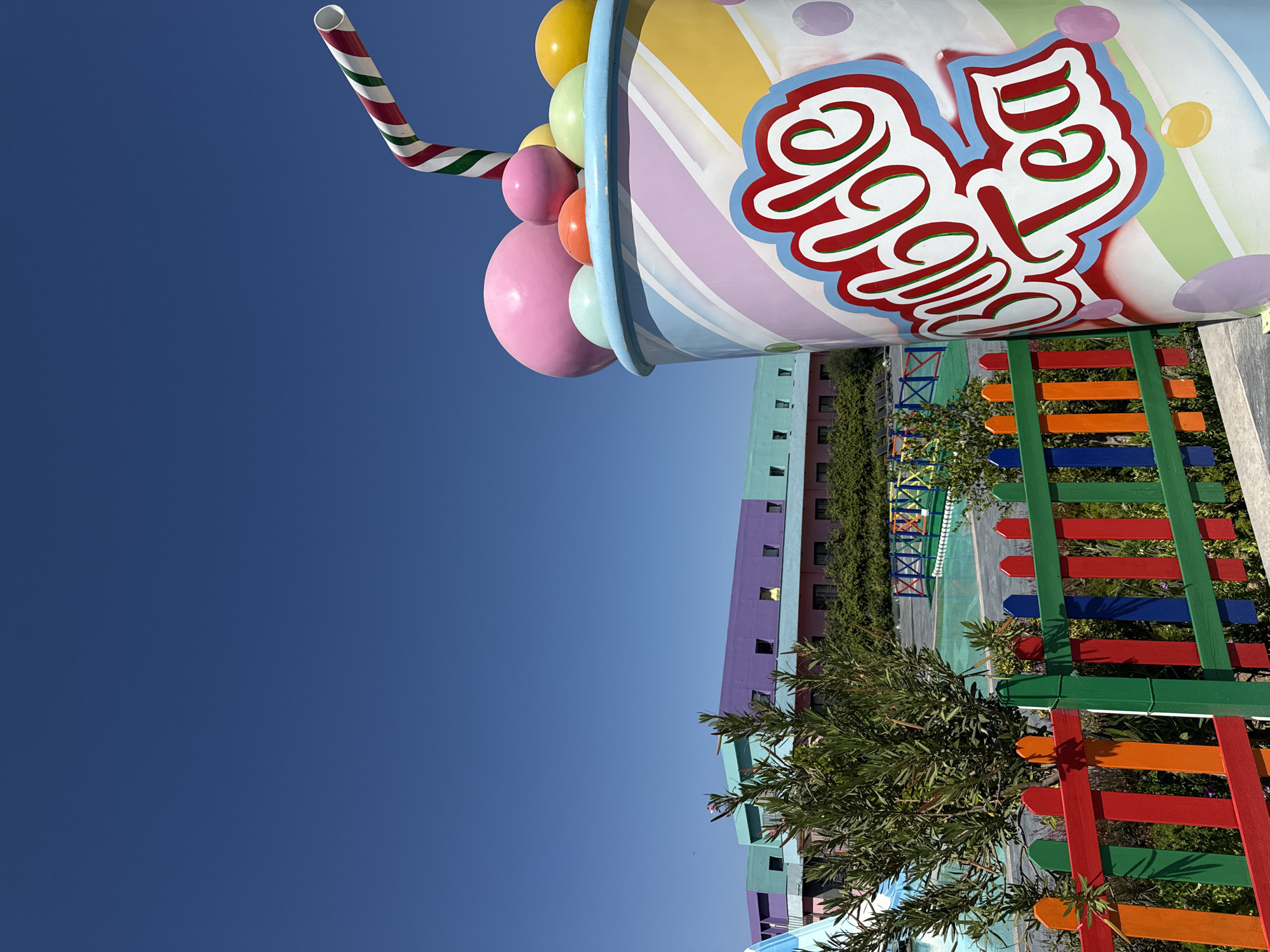 An outdoor shot shows a giant cup of bubble tea as a prop, with a vibrant, multi-colored building in the background. The building has sections painted in purple, turquoise, and pink. In front of the building, a swimming pool is visible, bordered by a colorful fence made of red, orange, green, and blue wooden pickets. A lush green tree with pink flowers stands to the left of the pool. The sky is a clear, bright blue. This scene is likely located at a resort or entertainment venue in Bodrum, Turkey.