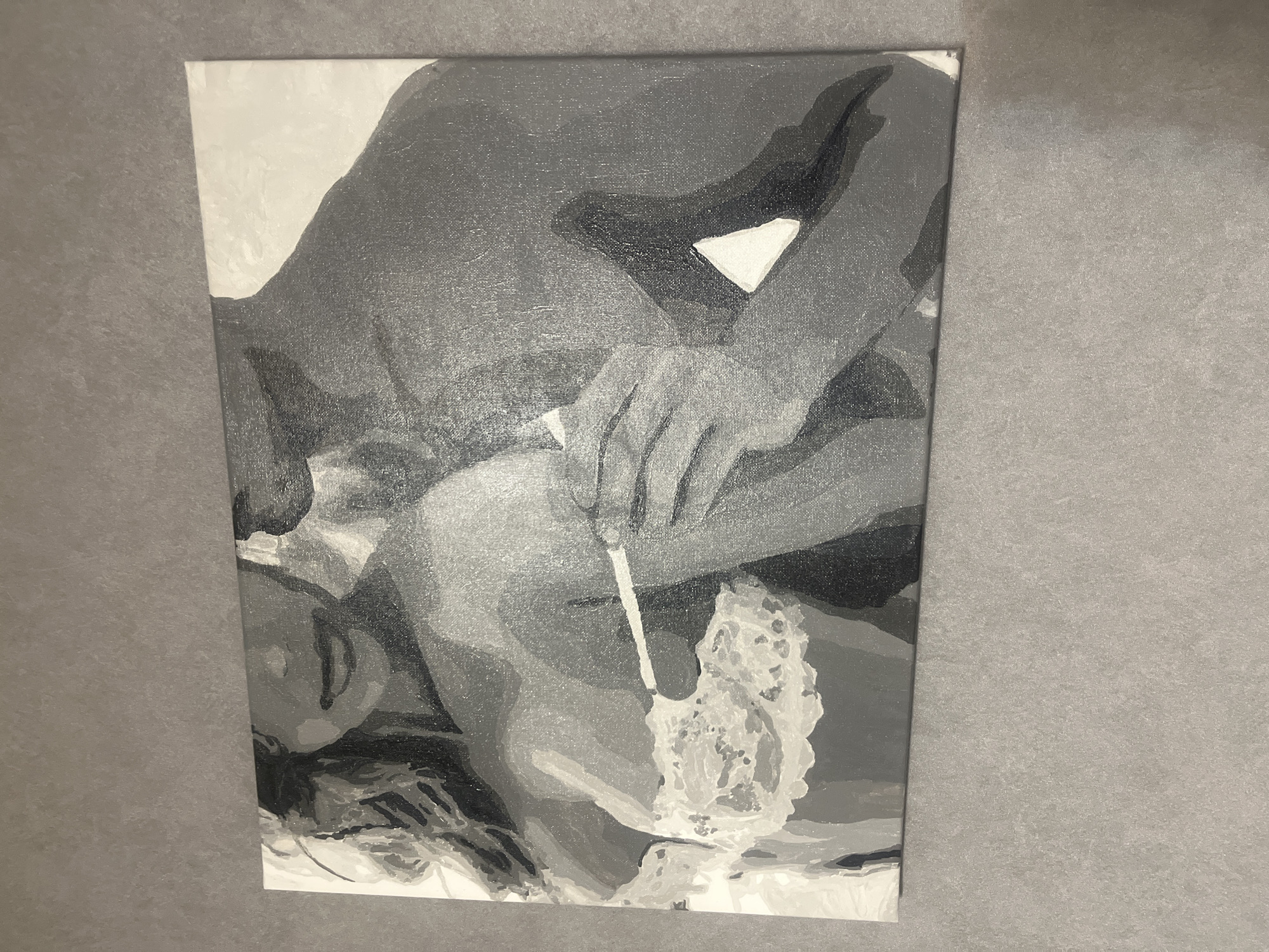 A monochrome painting on a canvas depicts a close-up of two individuals, a woman and a man, in an intimate pose. The woman, on the left, is wearing a lacy brassiere, her hair is wavy and extends down her left side. The man is on the right, his torso bare. His right arm is around the woman's back, and his hand is gently touching her shoulder and chest area, near her brassiere. Their faces are not fully visible, but their expressions appear soft. The painting uses a limited palette of greys and white, with distinct blocks of color suggesting a stylized or abstract representation of the figures. The canvas is hung on a textured grey wall. The overall impression is one of tenderness and connection. The specific location in Rybnitsa, Moldova is not ascertainable from the image.