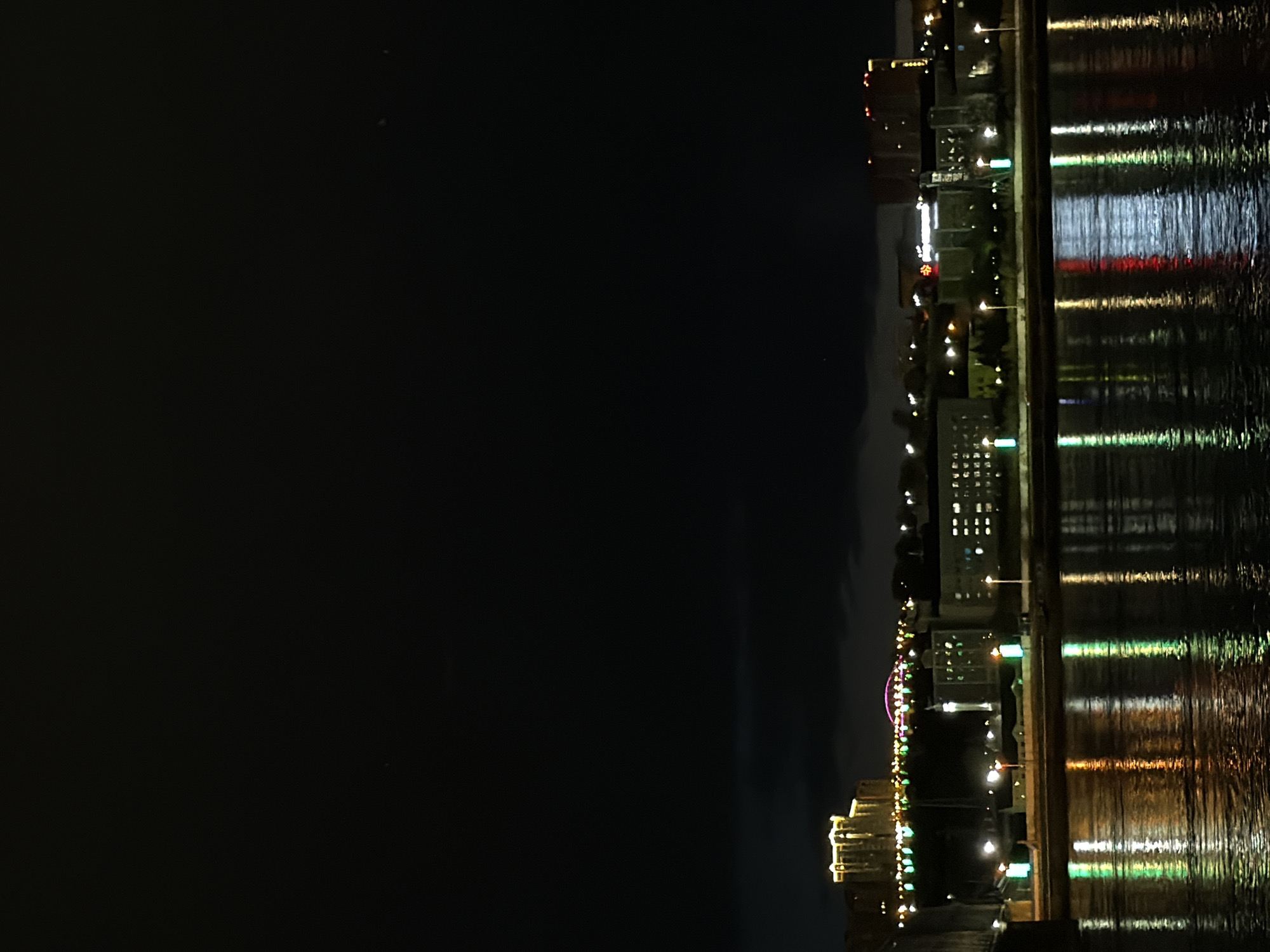 The image depicts the night skyline of Krasnoyarsk, Russia, reflected in the Yenisey River. The skyline is illuminated with various lights from buildings, streets, and other structures. The river's surface reflects these lights, creating a shimmering effect. The sky above is dark. The cityscape includes a Ferris wheel and several high-rise buildings.