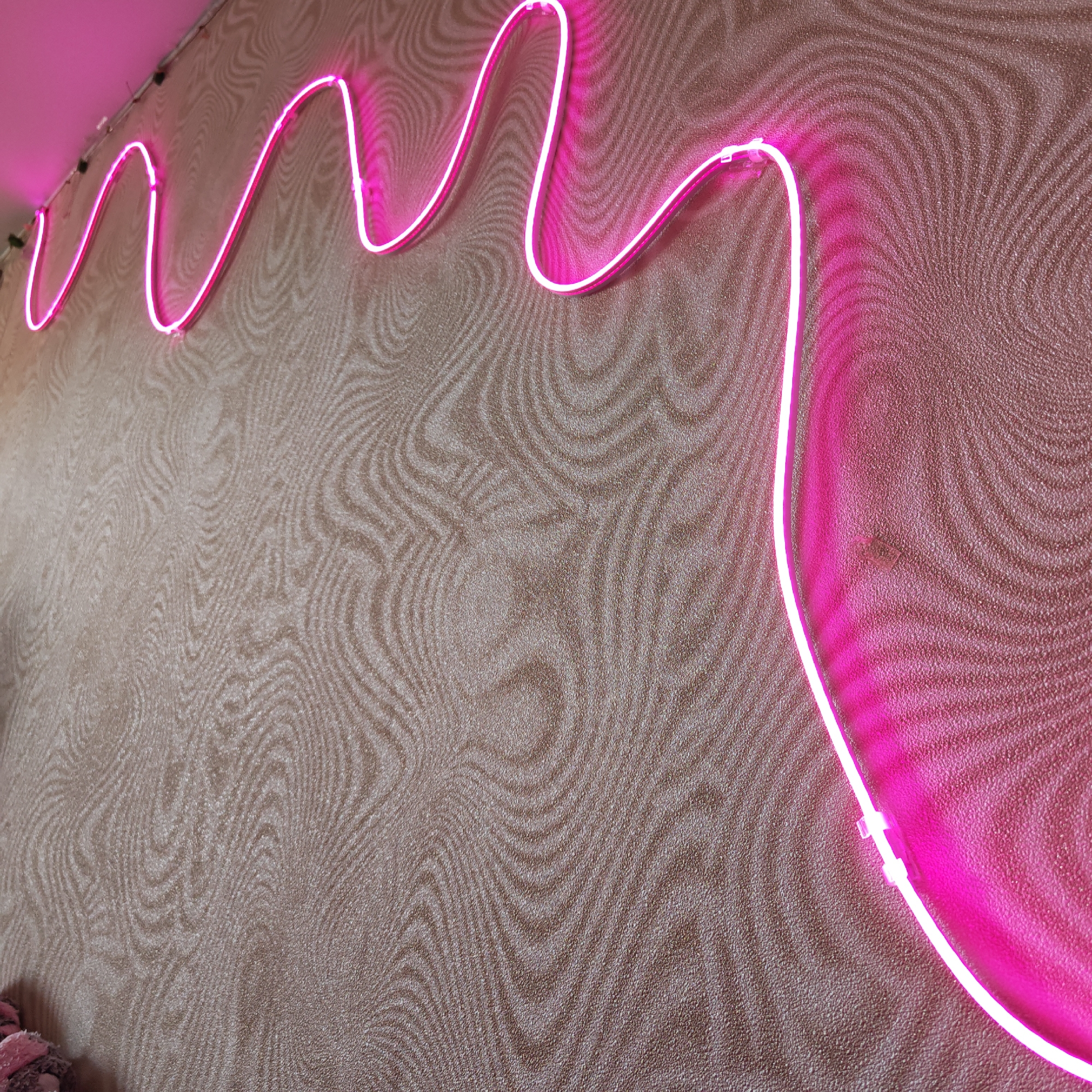 A low-angle shot shows a wavy pink neon light strip mounted on a textured wall. The wall has a repeating, swirling pattern in shades of brown and tan. The neon light strip starts at the top left and meanders across the wall, ending at the bottom right. The strip is attached to the wall with small, clear clips. The lighting casts a pink hue on the wall near the light strip. The image was taken in Rostov, Russia.