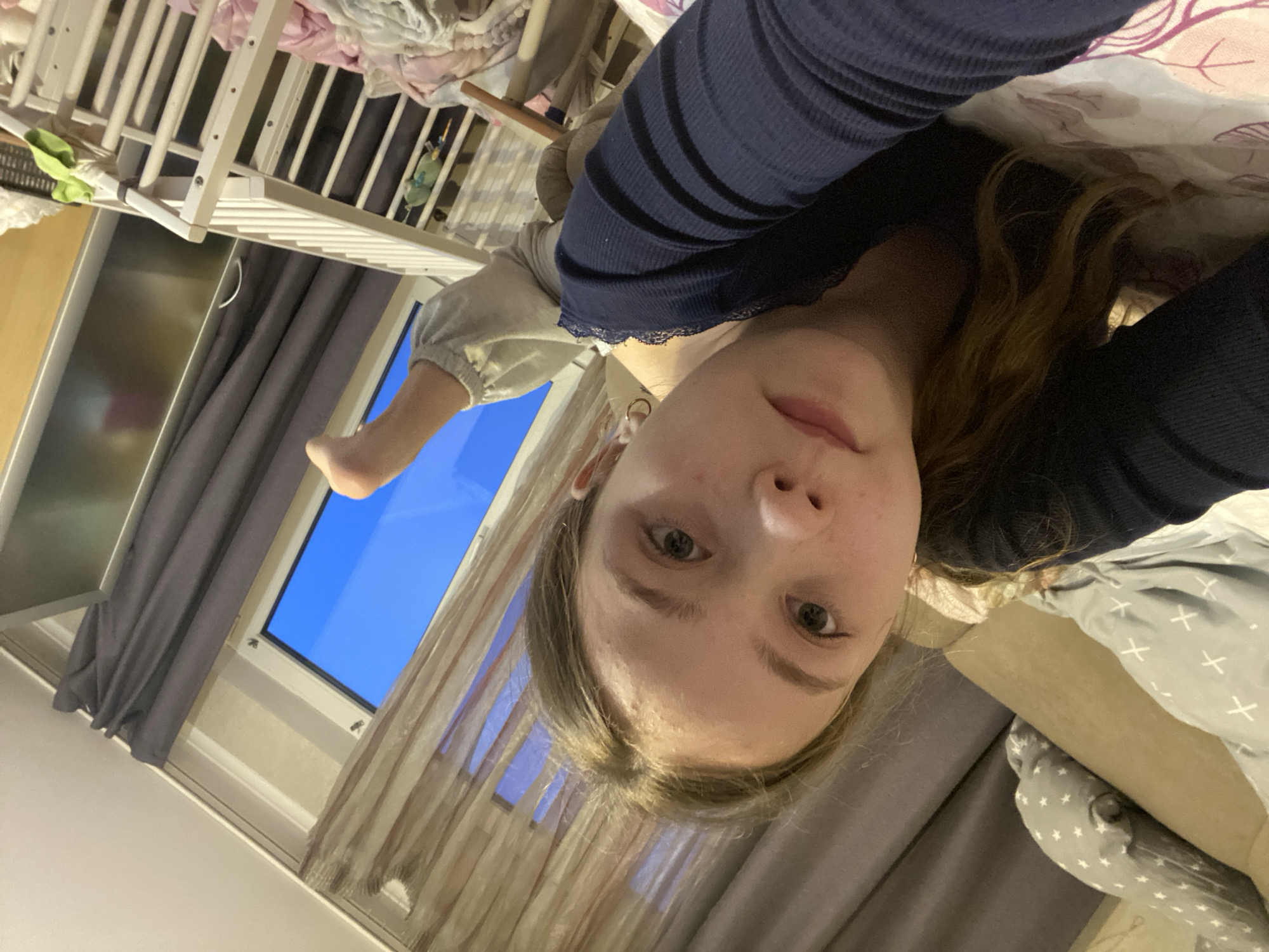 The media file captures a selfie taken indoors, likely in a bedroom or nursery. The image is rotated, showing a young person lying down, looking directly at the camera.
**Subjects:** The primary subject is a young person, seemingly female, with fair skin, light-colored eyes, and light brown or blonde hair. They are wearing a dark blue long-sleeved top with thin horizontal stripes and light grey or beige trousers. Their face is in the foreground, with a neutral expression. A bare foot, possibly belonging to the same person, is visible in the mid-ground towards the left. In the background, to the left of the person, is a white wooden baby crib containing crumpled fabric items, including pink and white textiles, and a green soft toy hanging on the bars. To the right of the person, a grey blanket with white cross patterns and a grey pillow with white star patterns indicate a bed.
**Scene and Setting:** The setting is an indoor room, consistent with a home environment. A window dominates the background behind the person's foot, revealing a clear, bright blue sky, suggesting daytime. Dark grey curtains hang on the left side of the window, while lighter, sheer curtains are on the right. A metal handle, possibly from a cabinet or refrigerator, is visible near the left curtain.
**Activity:** The person is taking a selfie from a supine position, likely while relaxing in bed.
**Notable Details:** The bright blue sky indicates it is daytime with clear weather. The presence of both a crib and a bed suggests a shared bedroom or a nursery. No visible text is present in the image. The overall mood appears calm and relaxed.