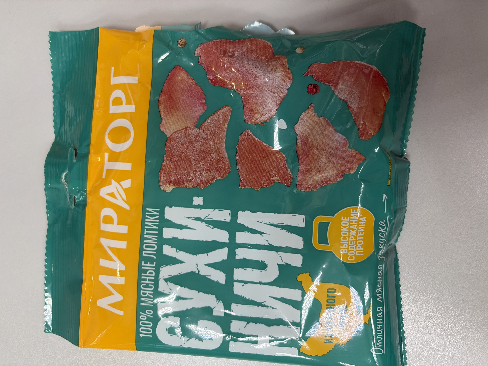 A teal and yellow flexible packaging of a food product, likely a snack, is centrally displayed on a light, textured surface. The package is slightly rotated, giving it a diagonal appearance. The main body of the package is teal, featuring multiple irregularly shaped, reddish-brown pieces of what appear to be dried meat slices. A prominent vertical yellow strip runs along the left side of the package.
Visible text in Russian identifies the product and brand. The brand name "МИРАТОРГ" (MIRATORG) is printed vertically in white on the yellow strip. Below this, it reads "100% МЯСНЫЕ ЛОМТИКИ" (100% MEAT SLICES). Large white text on the teal background clearly states "СУХИЕ ЛОМТИКИ" (DRIED SLICES). A graphic of a stylized turkey or chicken is present, with the text "ИЗ ИНДЕЙКИ" (FROM TURKEY) next to it, indicating the type of meat.
Towards the right side, a yellow badge shaped like a kettlebell contains the text "ВЫСОКОЕ СОДЕРЖАНИЕ ПРОТЕИНА" (HIGH PROTEIN CONTENT). Along the bottom right, there is handwritten-style text that says "Отличная мясная закуска" (Excellent meat snack), and in smaller print, "Вариант сервировки" (Serving suggestion). The setting appears to be indoors, possibly on a table. The overall impression is a commercial product, likely from Russia, given the language and provided location context of Sochi.