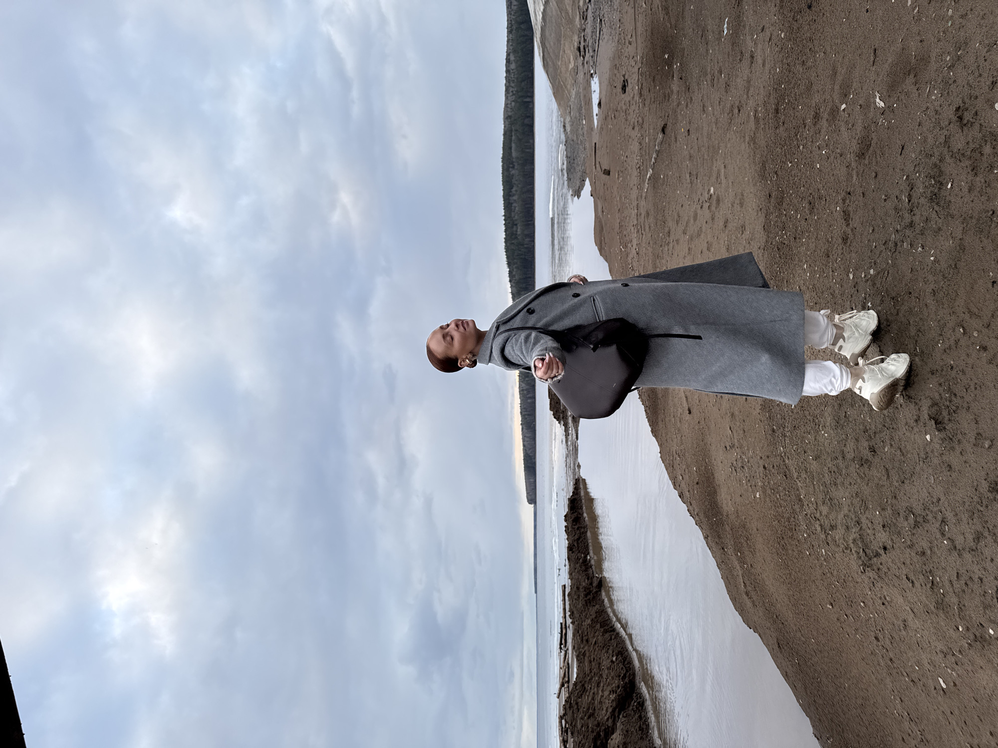An individual is captured standing on a sandy or muddy bank beside a wide body of water under an overcast sky in Chaikovsky, Russia. The person, with short reddish-brown hair, wears a long, double-breasted grey coat, white trousers, and light-colored chunky sneakers. A black bag is slung across their chest. Their head is slightly tilted upwards, eyes closed or looking towards the sky, conveying a sense of serenity or contemplation.
The scene suggests a cold environment, evidenced by the heavy coat and possible ice patches along the water's edge. The body of water, likely a river or lake, extends into the distance, with a dark, dense forest line visible on the far bank. The immediate foreground is composed of damp, dark brown earth, possibly a mix of mud and sand. The sky is uniformly covered with light grey clouds, indicating an overcast daytime, contributing to a soft, diffused lighting throughout the landscape. There is no visible text present in the image.