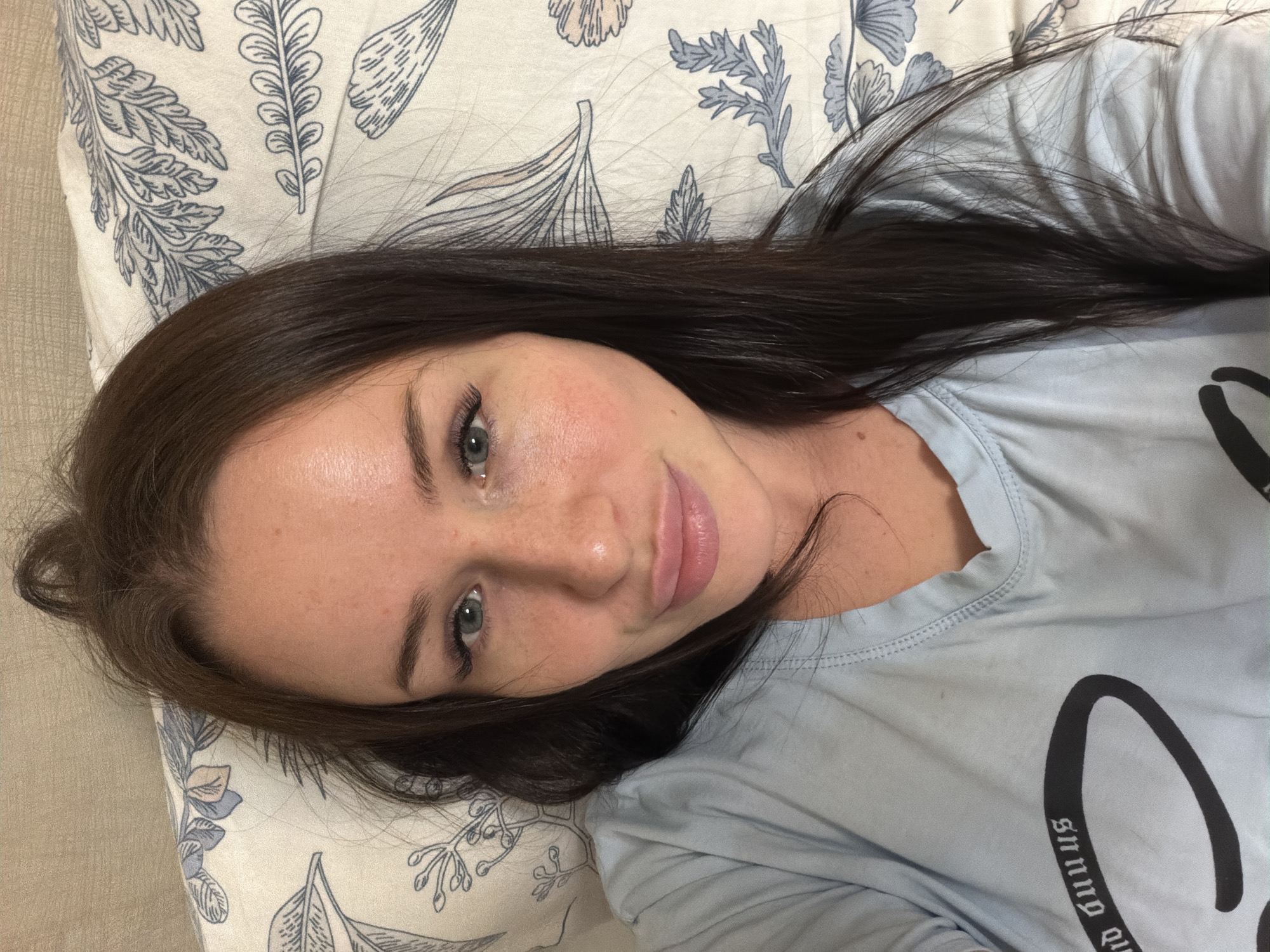 A young woman with long, dark brown hair and blue eyes is depicted lying down, looking directly at the camera with a relaxed expression and a slight smile. Her skin is fair, with visible freckles across her nose and cheeks. She is wearing a light blue t-shirt, on which partial black text is visible, appearing upside down and possibly mirrored, reading "Snnuq qu".
She is resting on a pillow that features a patterned design of blue and gray leaves and floral elements on a cream-colored background. Her hair is spread out on the pillow and around her head. The setting appears to be indoors, suggesting a comfortable and private space like a bedroom or living area. The lighting is soft and even, without strong shadows, indicating an indoor light source or indirect natural light. The overall mood of the image is calm and serene. The context provided indicates the location is Saint Petersburg, Russia, though no specific visual cues in the image confirm this.