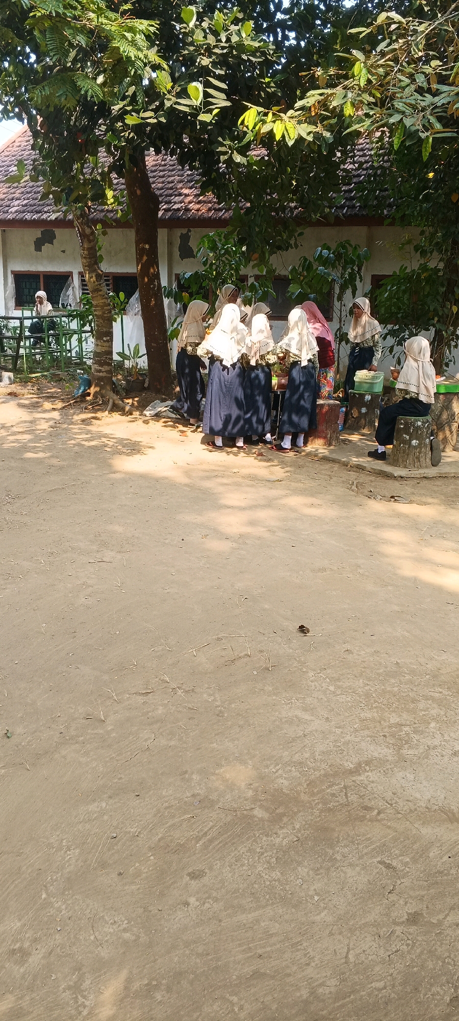 The image depicts an outdoor scene in front of a building with a tiled roof. Several young individuals wearing uniforms consisting of hijabs and dark skirts are gathered. Some are standing around a table, seemingly interacting with items on it. One individual is seated on a tree stump to the right, facing away from the viewer. A green metal structure is visible to the left, with one person seated on it. The ground is mostly dirt. The environment suggests a school or community setting in Penanggungan, Indonesia.