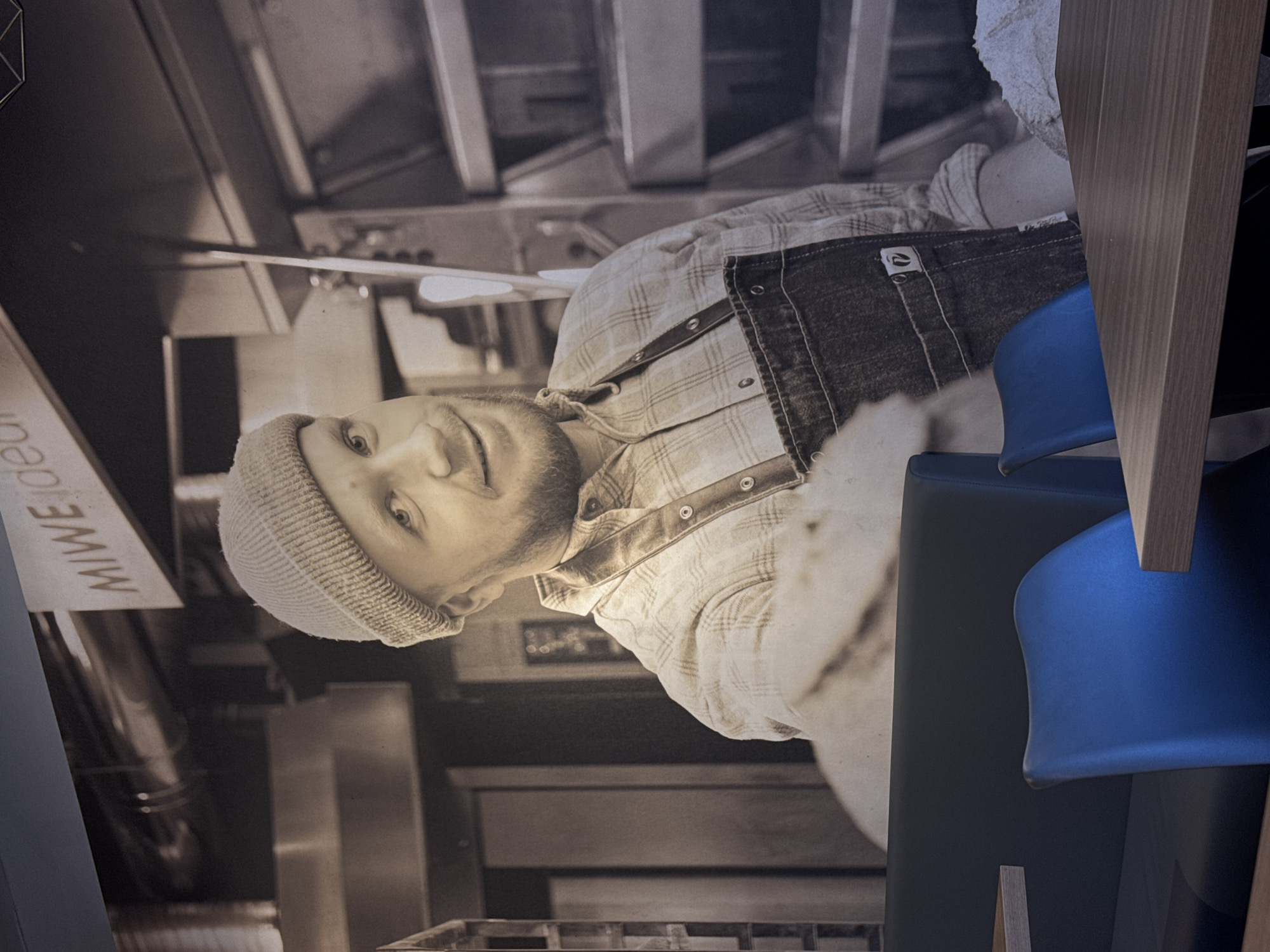 The image shows a monochromatic mural of a man wearing a beanie, a plaid shirt, and an apron. The mural is mounted on a wall near a MIWE logo and seems to be in the vicinity of some cooking equipment, hinting at a food-related business or establishment. A chair with a blue seat is in the foreground. The location is Saint Petersburg, Russia.
