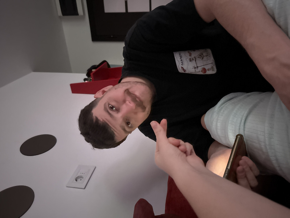 A man wearing a black t-shirt is turned sideways, looking at the camera with a slight smirk. A hand in the foreground forms a finger heart. In the background is a white wall featuring two brown, circular shapes and a white electrical outlet. A red object sits atop the surface near the wall. The man appears to be holding a phone and is partially covered with a light blue blanket.