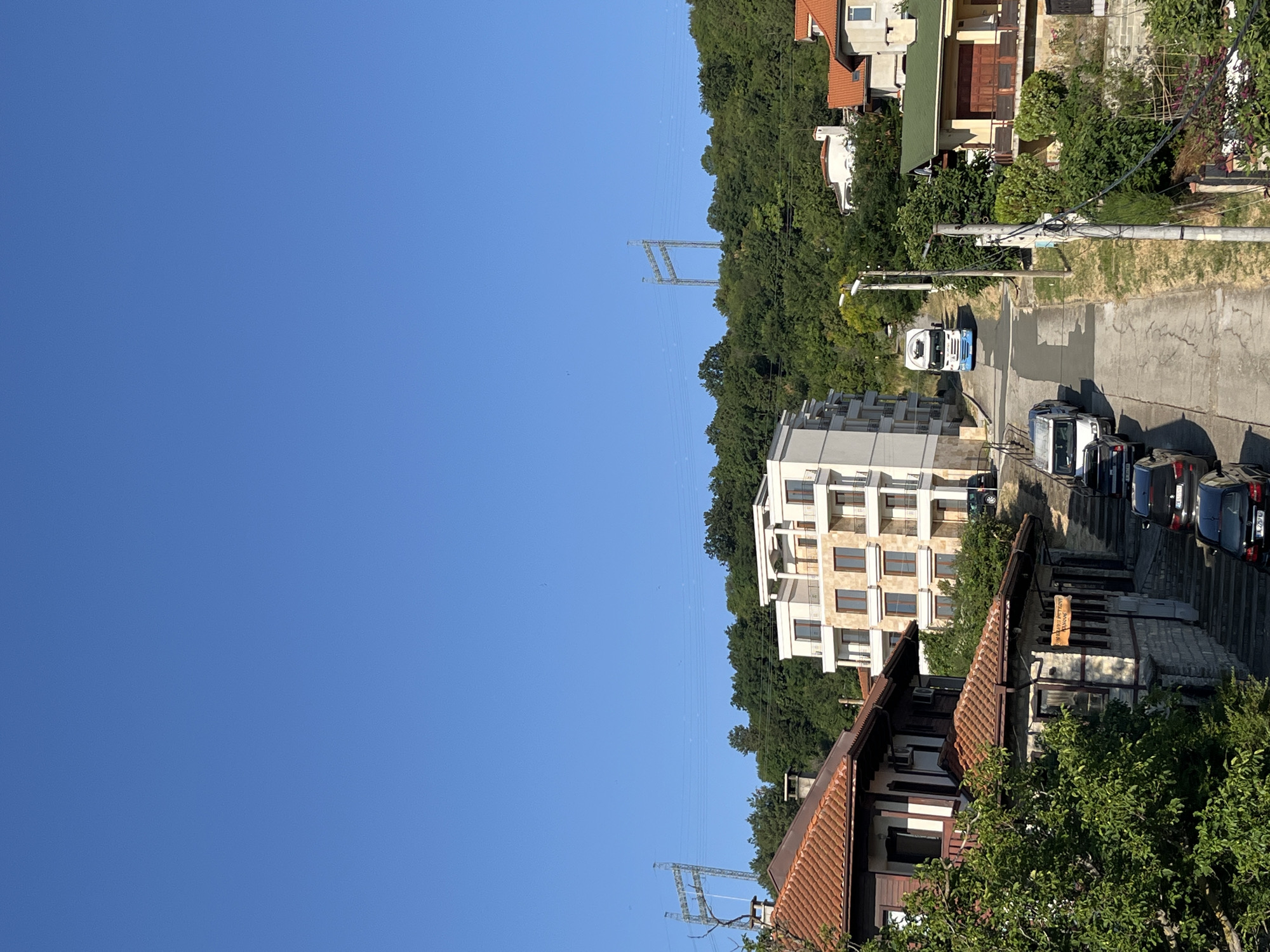 This is an eye-level, medium shot taken on a clear, sunny day in the city of Obzor, Bulgaria. The scene depicts a residential street winding uphill, lined with houses and a modern apartment building.
On the left side of the street, a traditional, two-story house with a red tiled roof is visible. Its lower story is made of stone, and the upper story is wooden with white accents. Next to it, a more modern, multi-story apartment building features a white facade with multiple balconies and numerous windows.
The street itself is paved and slopes upwards. A white truck with a blue stripe is seen driving up the hill. Several cars are parked along the sides of the street.
In the background, a lush, green hillside is covered with trees. Two tall, industrial power line towers are visible against the clear blue sky, situated on the hilltop. The overall impression is of a sun-drenched, hilly town with a mix of traditional and contemporary architecture.