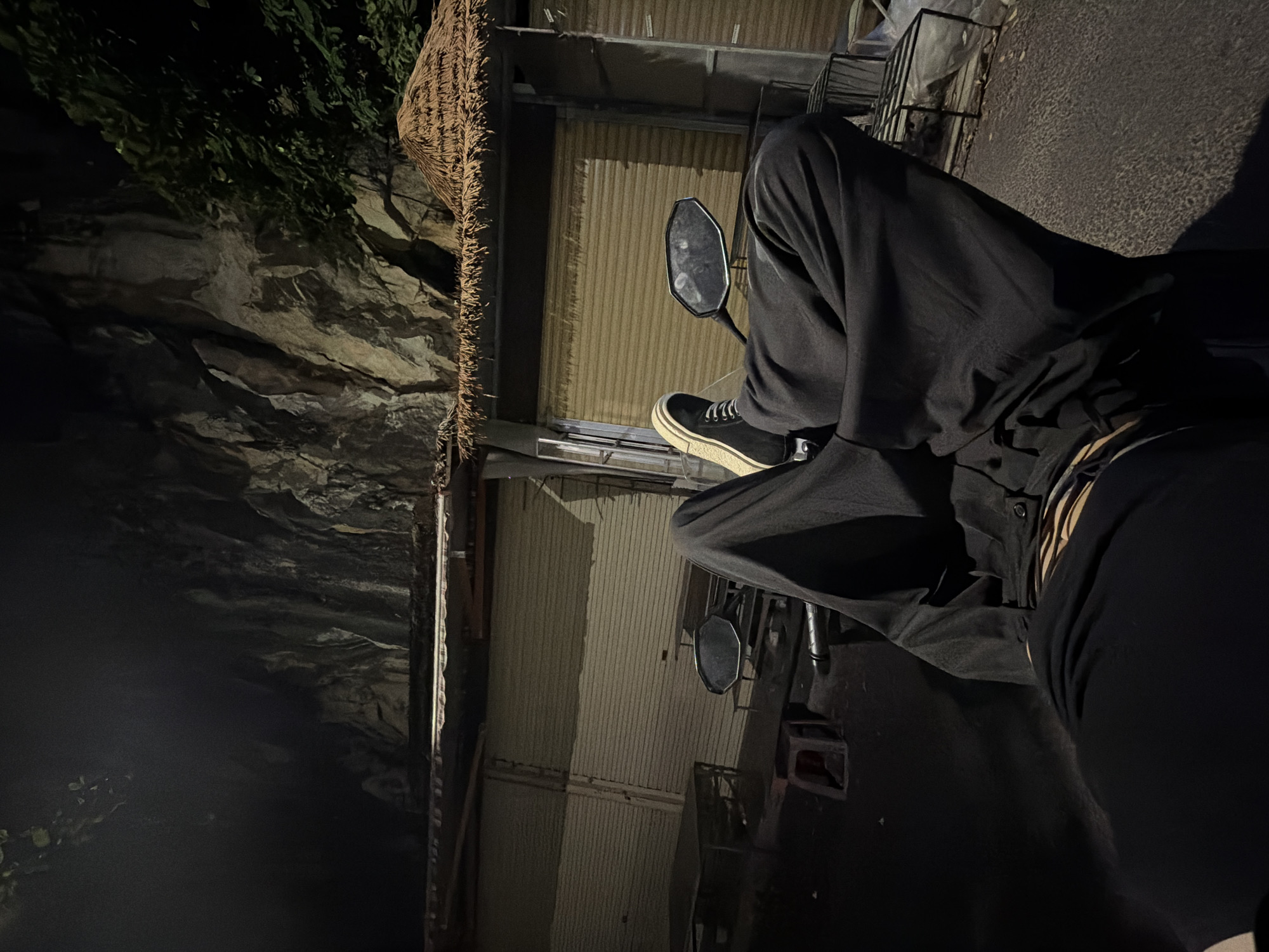 The image captures a nighttime scene featuring a person seated on a motorcycle or scooter in what appears to be an urban or semi-urban environment. The person's lower body is prominent, clad in black pants and black and white sneakers, with their legs positioned as if resting on the vehicle's footrests. A side mirror of the motorcycle is visible in the center-left, reflecting a blurred view.
The setting reveals diverse textures and structures under artificial lighting. To the right of the person, a tall corrugated metal wall or shutter, possibly part of a building, stands vertically, displaying its ridged surface. Adjacent to this, a rough, woven, or thatched element, possibly a roof edge or a decorative feature, is partially visible. To the left, a natural or extensively textured rocky wall, interspersed with dark green vegetation at its upper edge, creates a rugged backdrop. The ground surface beneath the motorcycle is dark and textured, suggestive of asphalt or a paved area.
The overall lighting is dim, characterized by deep shadows and highlights that accentuate the textures of the surroundings. The time of day is clearly night, with light sources external to the scene casting strong contrasts. There is no discernible activity beyond the person being seated on the vehicle, possibly waiting or parked. No visible text is present in the image. The scene's character is consistent with the provided location context of Đà Nẵng, Vietnam.