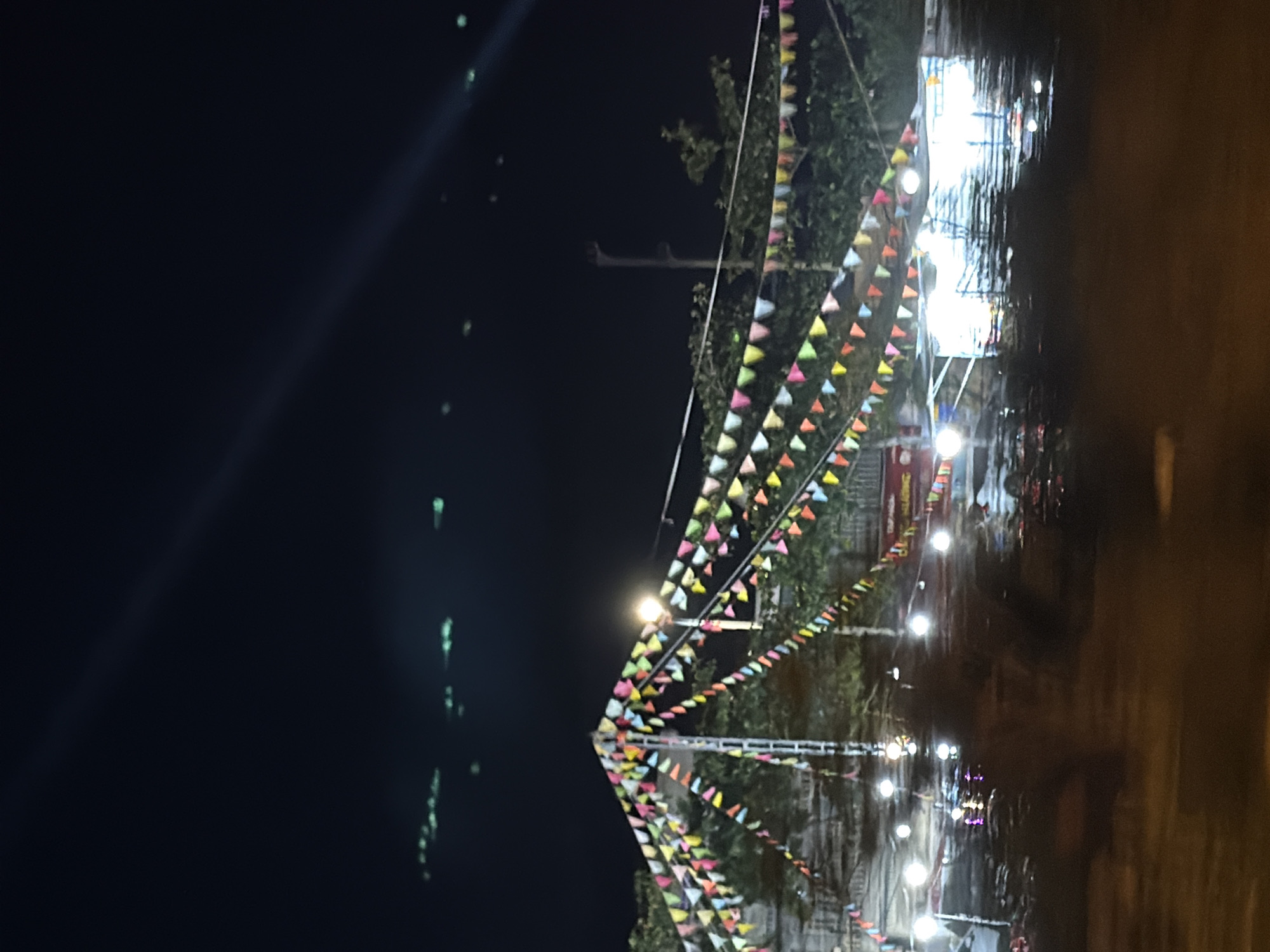 In Nha Trang, Vietnam, a night scene unfolds under a dark sky, illuminated by various light sources. The foreground features an out-of-focus, blurred view of a street, possibly indicating motion blur from a vehicle. In the mid-ground, a festive atmosphere is evident with strings of colorful triangular flags stretching across the frame, strung between poles and trees. These flags are a mix of yellow, pink, green, blue, and white, creating a vibrant display. A bright light fixture is visible atop one of the poles, casting a warm glow. Behind the flags, a market or food stall area is suggested by structures covered by translucent sheeting, reflecting the numerous bright lights illuminating the space. Several round, bright lights are scattered throughout this area, some appearing to be suspended. A banner with text, partially obscured, is visible on the right side. The background is dominated by the dark night sky, with a faint diagonal light streak stretching from the upper right to the mid-left. In the distance, a cluster of small, blurry green lights are faintly visible, possibly distant city lights or reflections. The overall impression is of a lively outdoor event or marketplace at night.