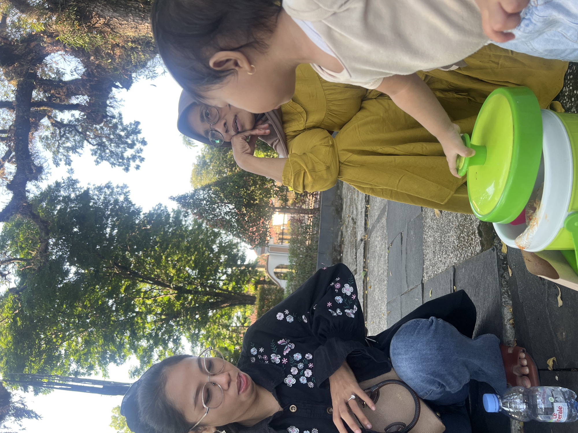 The image captures three individuals outdoors, likely in a park or garden setting in Purwodadi, Indonesia. The scene appears to be bathed in natural daylight, suggesting it was taken during the day.
In the foreground, a person is seated, wearing a black embroidered jacket and blue jeans. They have glasses on and are looking towards the right of the frame. Their hand rests on a brown bag, and a bottle of water with a blue cap is visible next to their foot.
To the left of the seated person, another individual is partially visible, wearing a hijab and glasses. They are looking upwards and to the left, with their hand resting on their chin.
In the background, a young child is interacting with a green and white plastic toy. The child is wearing a light-colored top and is reaching out to the toy. The older individuals are looking at the child.
The setting includes trees with lush green foliage, casting shadows on the ground. The ground itself is paved with rectangular stones. In the distance, hints of buildings with arched windows and red roofs are visible, suggesting a developed area nearby.
The overall mood seems relaxed and casual, possibly a family outing or a gathering of friends enjoying the outdoors. The interaction between the child and the adults suggests a moment of playful engagement. No specific text is clearly legible on the objects within the frame, beyond what appears to be a brand name on the water bottle.