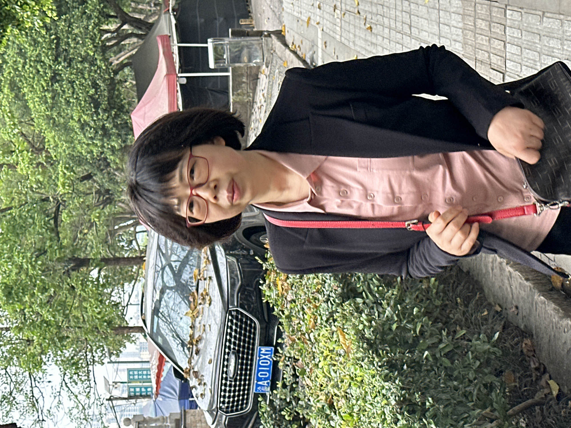 A person wearing red glasses, a light pink shirt, and a black blazer is standing outdoors. The person is holding a dark-colored bag with a red strap. Behind the person is a dark-colored car with leaves on the windshield. The car's license plate reads "A·010XM." The car is parked near a hedge of greenery. In the background are trees, a building with a red awning, and tiled sidewalk. The scene is likely Jiangjin, China based on the license plate and visual details of the environment.