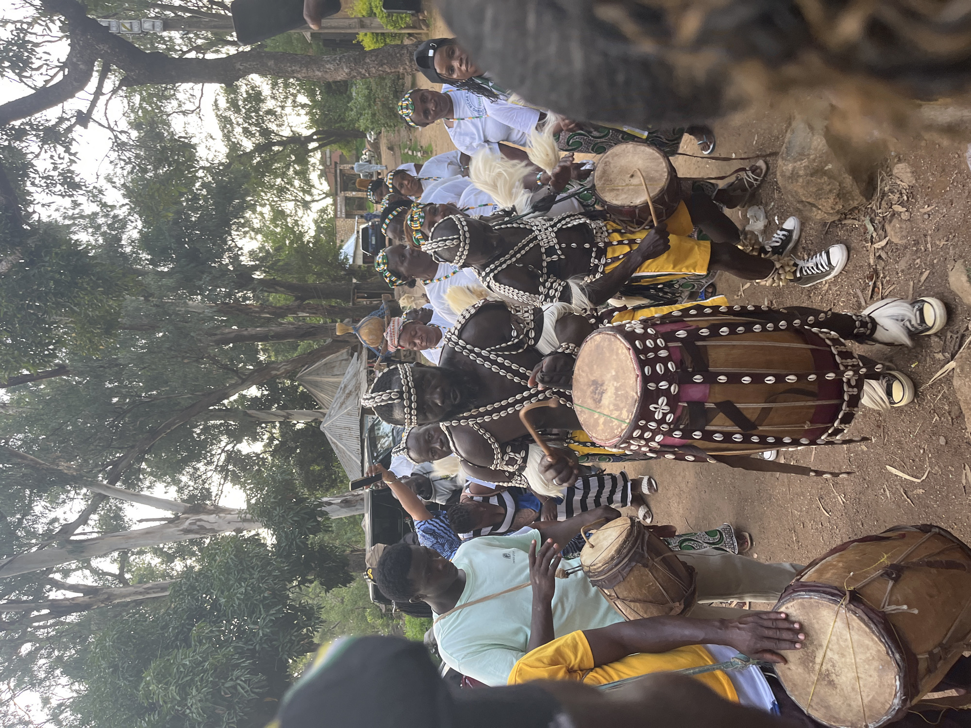 This media file depicts an outdoor gathering in Jos, Nigeria, likely a cultural event or performance. The primary subjects are a group of people, some of whom are dressed in traditional attire adorned with shells and beads. Several individuals are actively playing drums, holding drumsticks and striking the instruments. The drums themselves are varied in size and design, with some featuring intricate shell embellishments.
The scene is set amidst trees and earthy ground, suggesting a natural or semi-rural environment. The time of day appears to be daytime, with diffused sunlight filtering through the foliage. The overall atmosphere seems to be one of engagement and community. Some individuals are looking towards the drummers, while others are focused on their instruments. The clothing of some participants, particularly those playing the drums, is elaborate and ceremonial, indicating a significant event.
One drum is particularly detailed, wrapped in a dark fabric with rows of small shells and patterned designs. The players' hands are visible, actively engaged in drumming. In the background, more people are gathered, and there are hints of structures and vehicles, suggesting the event is taking place in a populated area. The overall impression is a vibrant display of cultural tradition and musical performance. No visible text is present in the media file.