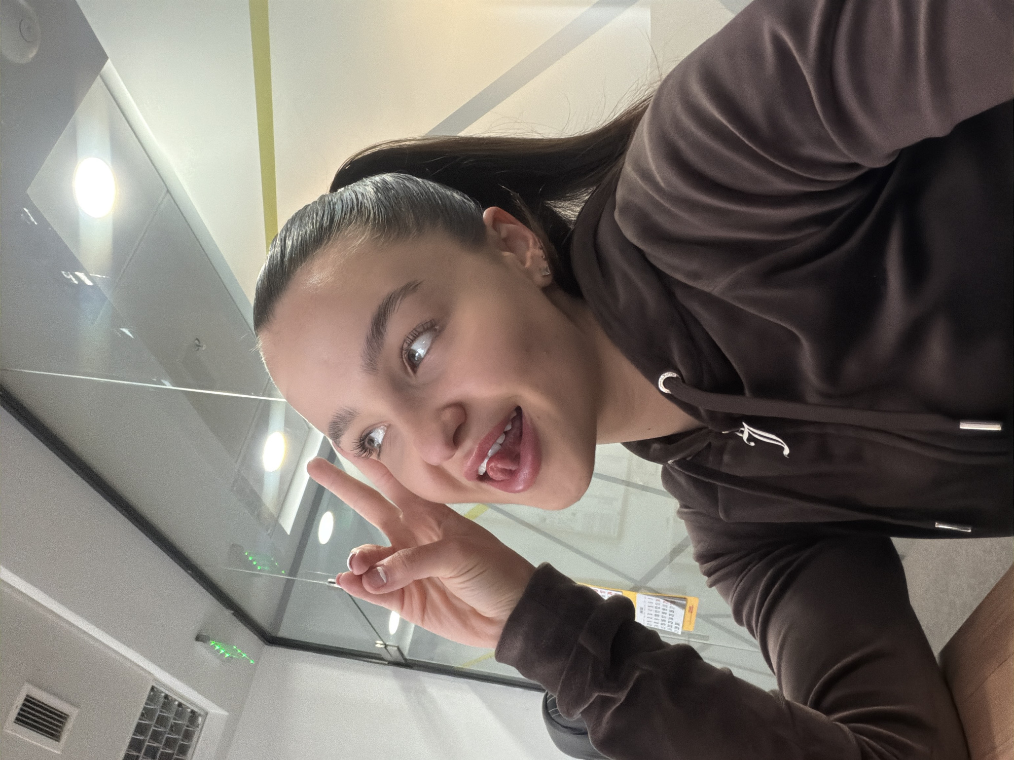 A young woman is pictured in a selfie. She has her hair pulled back in a ponytail and is wearing a brown hooded sweatshirt. She is making a peace sign with her right hand and sticking her tongue out. The background suggests an indoor environment, possibly a gym or recreational facility. The image is taken at an angle, looking upward at the woman. The exact location in Sofia, Bulgaria, cannot be determined based on the image alone.