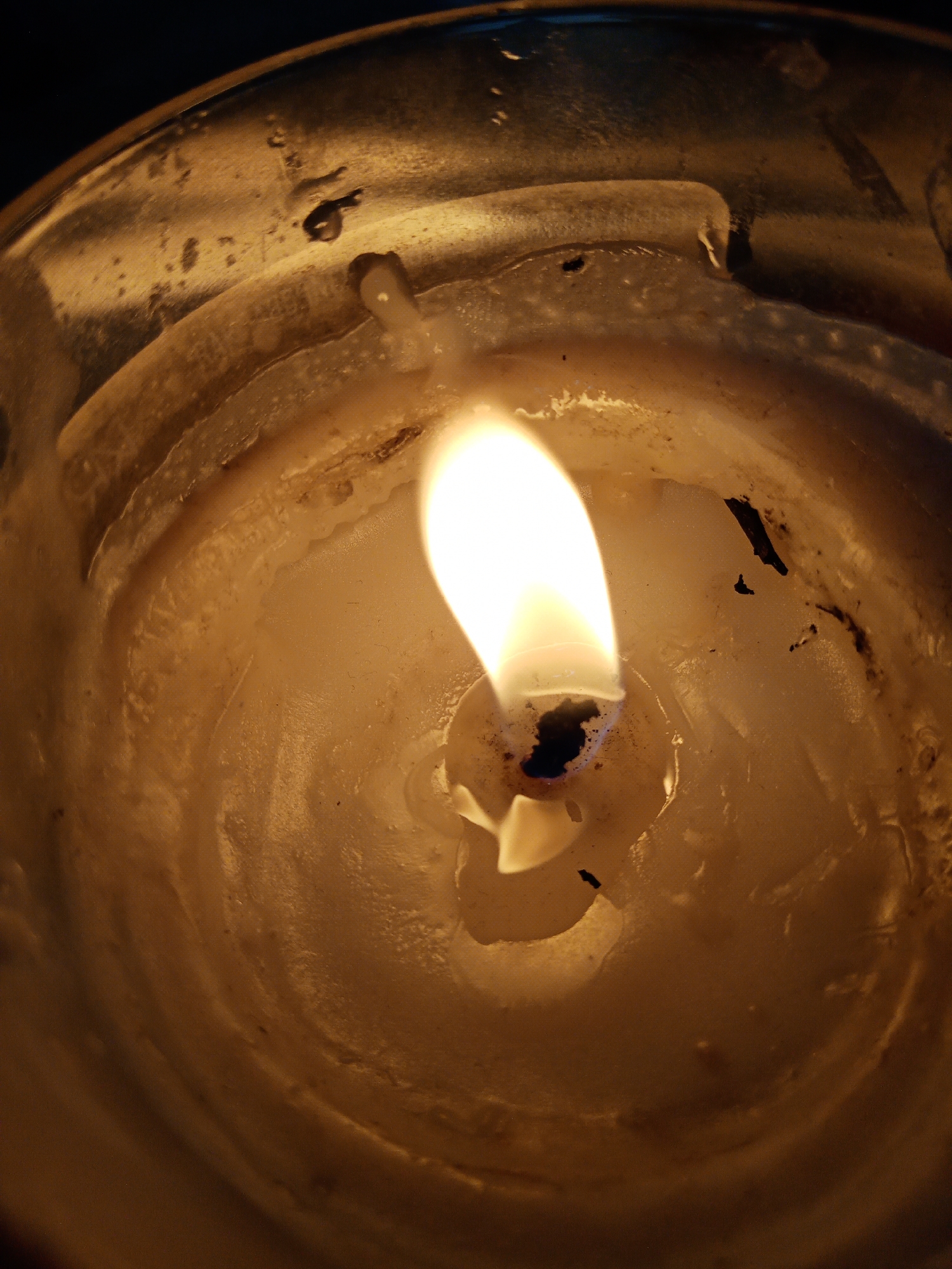 An intimate, top-down close-up captures a single burning candle, likely housed within a glass container, in Elektrostal, Russia. The primary subject is the vibrant, elongated flame, which glows with bright white and yellow hues, featuring a subtle blue base around the black, charred wick. The flame casts a warm, golden light across the surrounding melted wax, which forms a shallow pool around the wick and shows concentric rings and solidified drips along the inner walls of the container.
The wax itself is an off-white or cream color, with several dark specks and streaks, possibly from soot or debris, particularly near the wick and on the container's sides. The glass container appears to have accumulated layers of cooled, irregular wax, creating textured surfaces that reflect the candle's light. The background is dark, indicating a low-light environment, suggesting the image was taken during the evening or night. No people or visible text are present. The scene evokes a sense of calm and warmth.