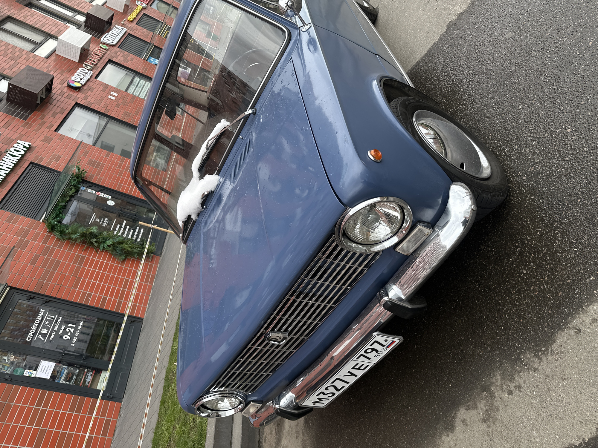 The image presents a slightly tilted view of a vintage-style blue car, likely a Lada, parked on an asphalt street in front of a red brick commercial building in Moscow, Russia. The car, occupying the foreground and middle ground, features prominent chrome bumpers, a chrome grille with an emblem, and round headlights. An orange side marker light is visible on the front fender, and patches of snow rest on the windshield wipers, indicating cold weather. The Russian license plate, "M327 YE797 RUS" (with region code 797 for Moscow and Moscow Oblast), is clearly displayed on the front. The vehicle has silver hubcaps, and the asphalt ground appears wet, suggesting recent precipitation or melting snow.
The background consists of a red brick building with multiple windows and several air conditioning units. Numerous Cyrillic signs on the building indicate various businesses. Visible text includes "ОПТИКА" (Optics/Optician), "ФОТО СРЕМОНТС" (Photo repair/services), "ФОТО НА ДОКУМЕНТЫ" (Photos for documents), "КСЕРОКОПИЯ И СКАН" (Photocopy and Scan), "ПЕЧАТЬ ФОТОГРАФИЙ" (Photo printing), and "СТРОЙХОЗМАГ" (Hardware store/Building materials store) with operating hours "ПН-ВС 9-21" and a phone number. Other services advertised include "НИКЮРА" (part of 'Маникюра' - manicure), "LPG массаж," "Услуги бровиста" (Brow artist services), and "тевой сервис" (likely 'ногтевой сервис' - nail service). An entrance to one of the shops, decorated with green garland, displays hours "10:00-22:00". The scene depicts a typical urban setting with commercial establishments, under what appears to be an overcast sky. No people are visible, and the car is stationary.