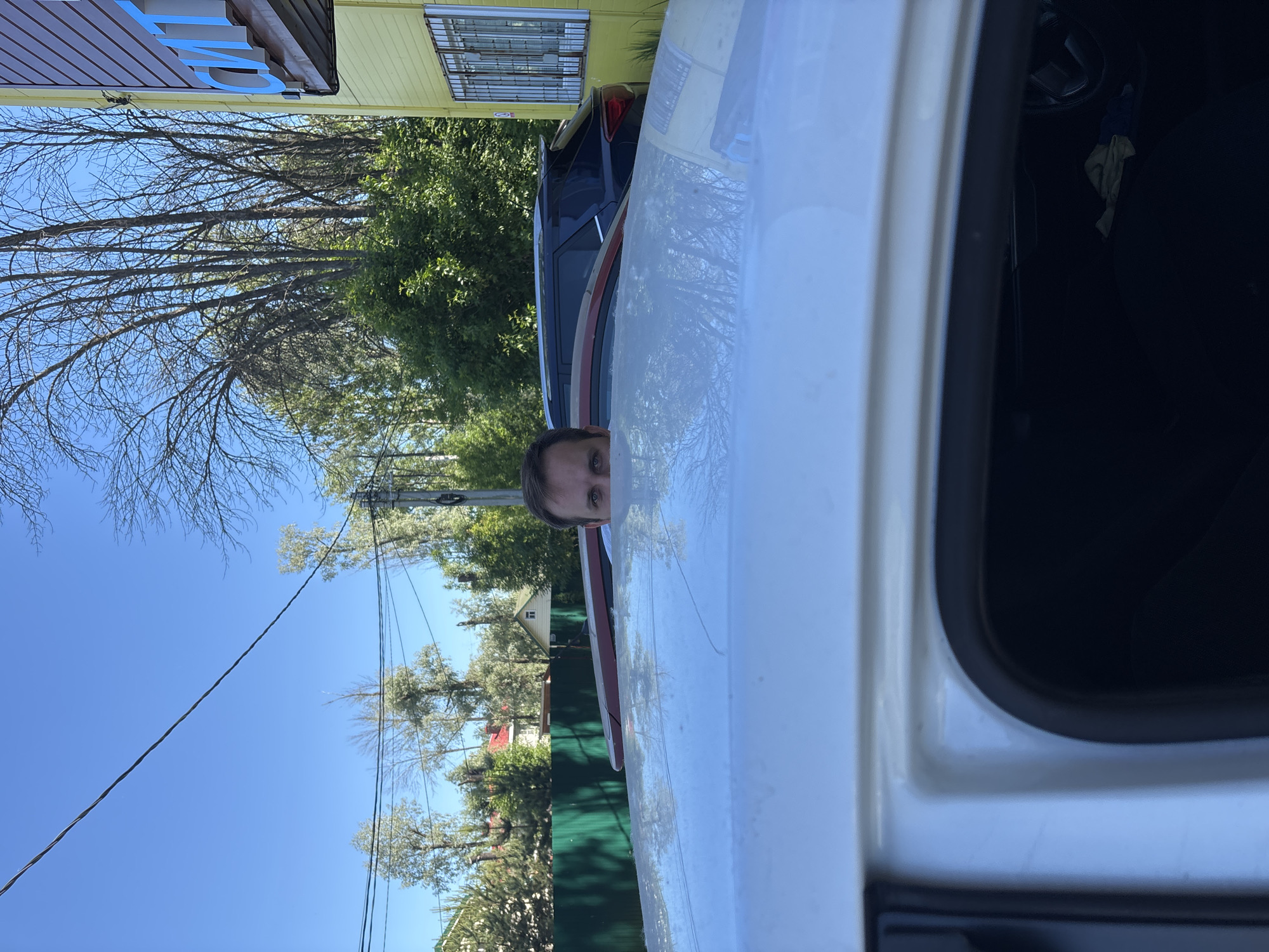 The scene depicts an outdoor environment in Rusanovo, Russia, under a clear blue sky. In the immediate foreground, the white roof and upper driver's side interior of a white vehicle are visible, with a steering wheel and a crumpled fabric item inside. Peeking over the white vehicle's roof, an adult male's face, specifically his eyes, nose, and forehead, is visible, looking directly towards the viewer. Behind the white vehicle, a red vehicle is partially visible, followed by a dark blue SUV. To the left, a dark green fence separates the immediate area from houses with red and white roofs amidst numerous trees. To the right, a two-story building with yellow siding features barred windows and a partial blue sign displaying the Cyrillic letters "СИТ". Various trees, some bare and some with green foliage, fill the background. Utility lines are visible across the sky.