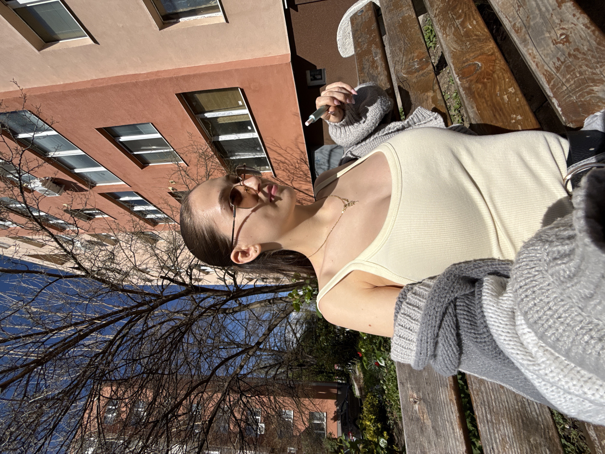 A young woman sits on a wooden bench in an outdoor setting in Sofia, Bulgaria, holding an e-cigarette. She is wearing sunglasses, a yellow tank top, and a gray sweater. A necklace is visible around her neck. In the background, there is a red-brick building with multiple windows and a bare tree. The weather appears to be sunny, as indicated by the shadows and brightness of the scene.
