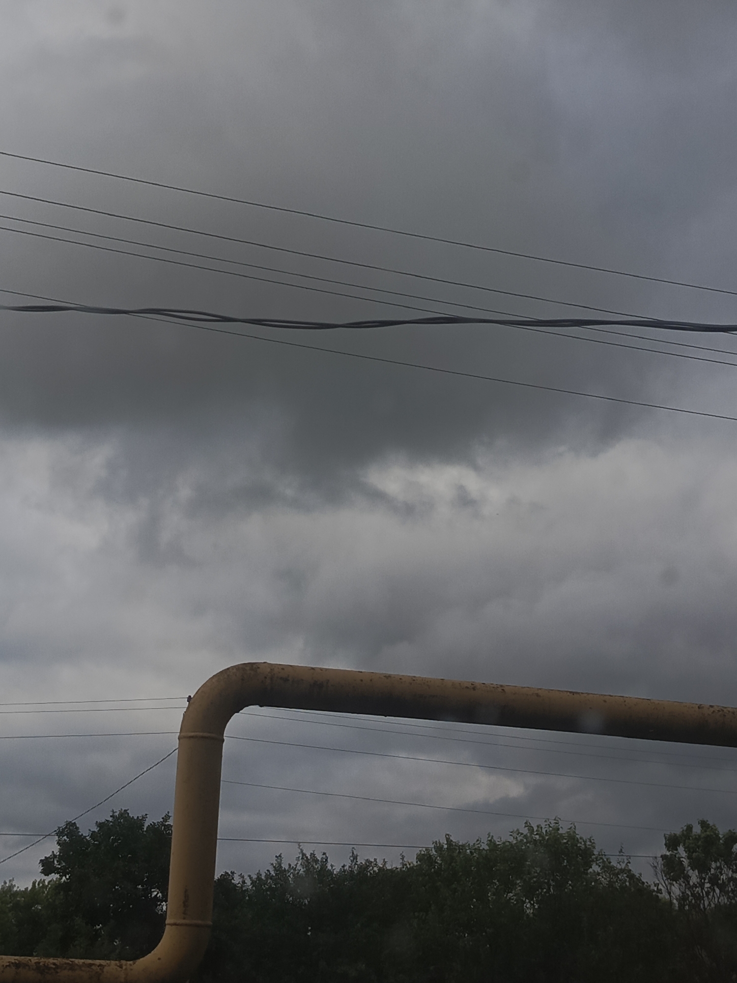 The image depicts an overcast sky with dark, heavy clouds. Several power lines run horizontally across the upper portion of the frame. In the middle ground, a large, yellow pipe, likely industrial, traverses the scene, with a section curving downwards. Below the pipe, a dense line of dark green trees forms the horizon. The overall setting appears to be an outdoor, possibly semi-industrial or utility area. There are no people or specific identifiable structures in the image, other than the general landscape elements. The scene could be located in Petrovka, Russia, given the context provided, but there are no direct visual cues to confirm this location.