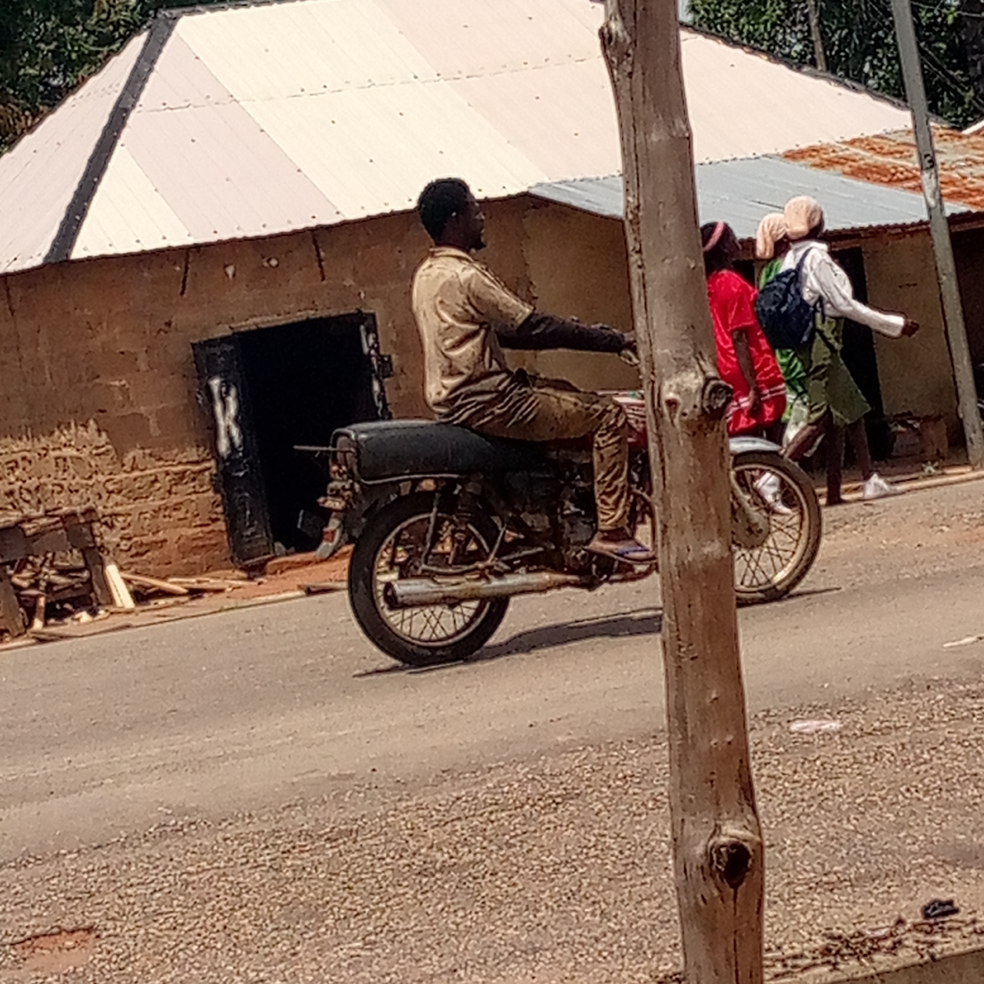The scene takes place in Mararaba, Nigeria. A man in a light-colored shirt and pants sits on a black motorcycle on a paved road. Behind him is a single-story building with a light-colored metal roof and a dark, possibly burnt-out doorway. To the right of the man, partially obscured by a tree trunk, are three people. One is wearing a red shirt, one is wearing a green skirt and white shirt with a black backpack, and the third is wearing a white shirt. The road appears slightly gravelly.