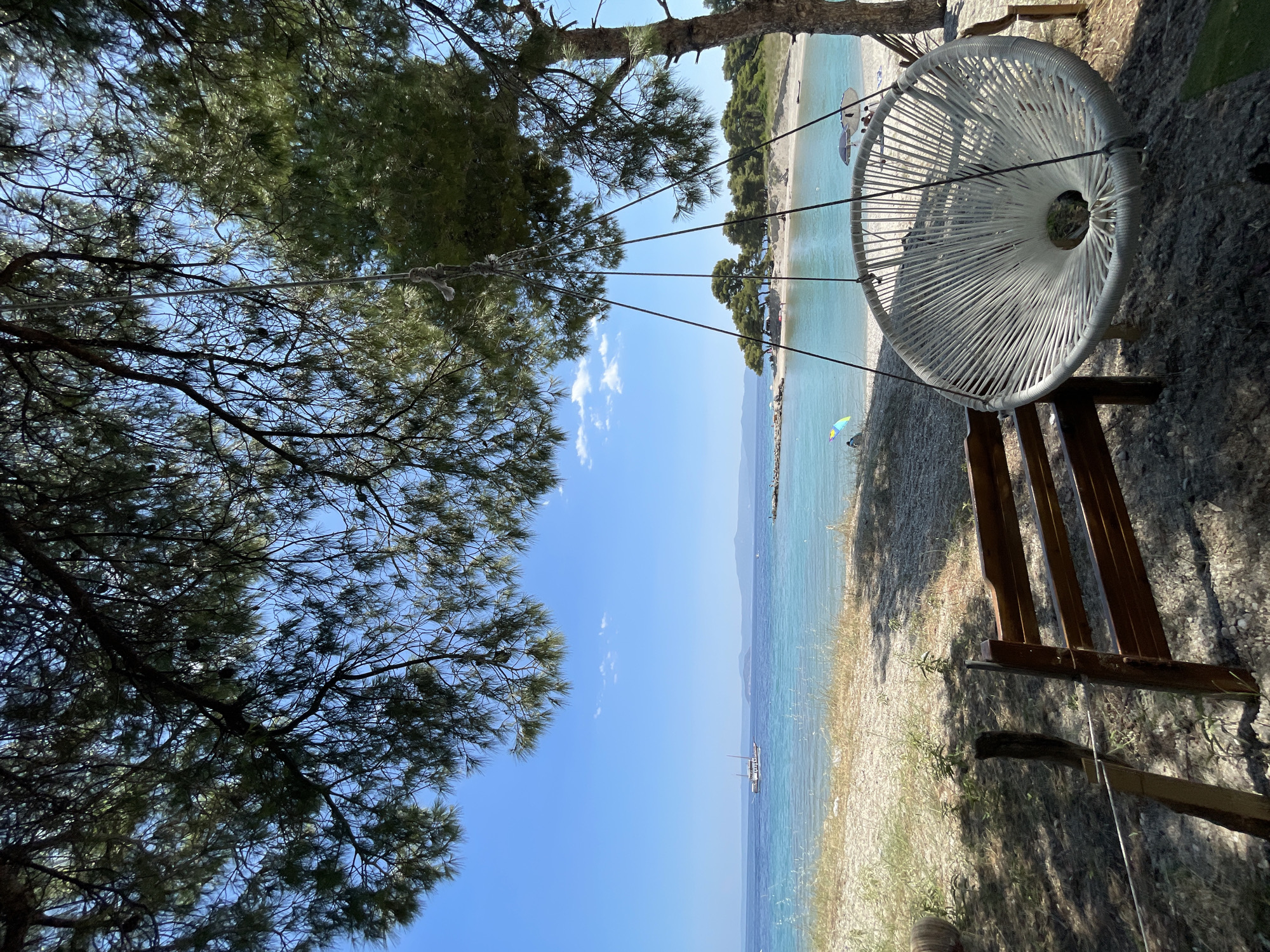 A white, circular, woven hanging chair is suspended from tree branches by ropes on a beach in the Municipal Unit of Pallini, Greece. The chair is in the foreground, to the right of a wooden bench. Behind the chair, the turquoise sea meets a sandy shore dotted with trees and a few people lounging on the beach. The sky is a clear, bright blue with a few scattered white clouds. A sailboat is visible on the horizon. The overall scene suggests a serene and relaxing day at the beach.