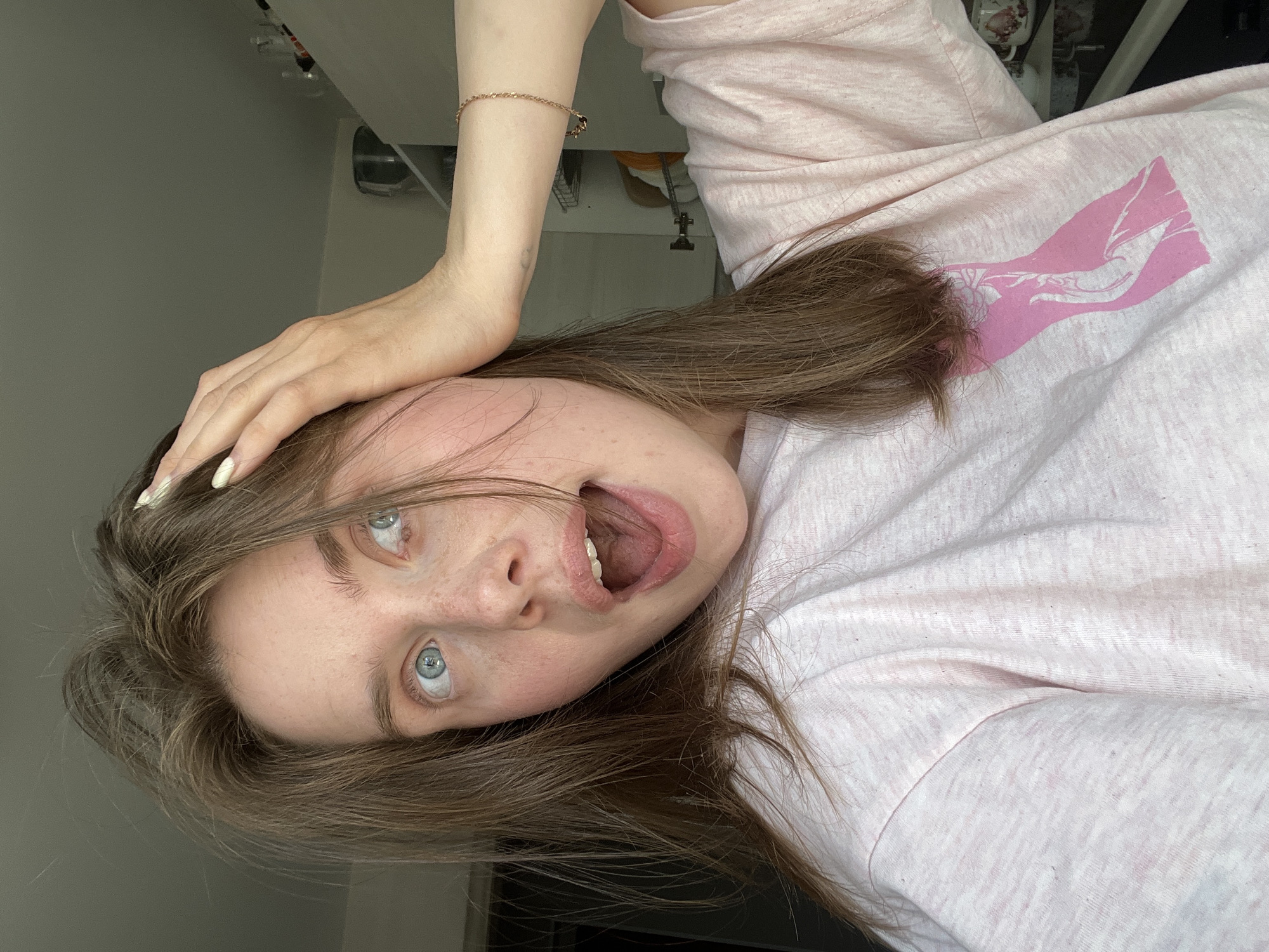 This is a medium close-up selfie of a young woman with light brown hair and blue eyes. She is wearing a pink t-shirt with a pink graphic design on it. Her right hand is raised and placed on her head, and her mouth is open in an expression of surprise or excitement. She has freckles on her cheeks and nose. The background is blurred and appears to be an indoor setting, possibly a kitchen with cabinets visible. There is no visible indication of the city of Chaikovsky, Russia.