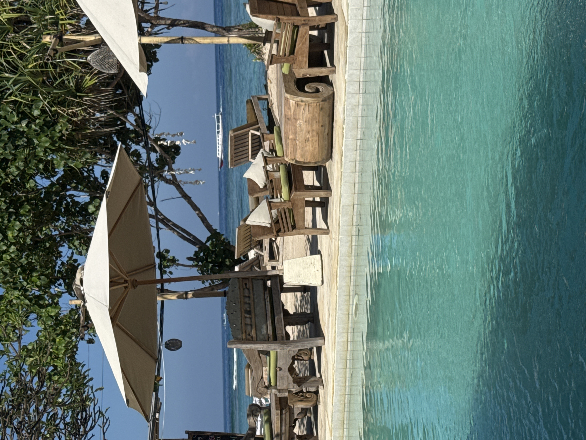 The image depicts an outdoor seating area adjacent to a swimming pool, likely in Unknown, Indonesia. Wooden chairs and tables are arranged along the pool's edge, shaded by a large parasol. The ocean is visible in the background with a small boat. The overall environment suggests a resort or vacation setting.