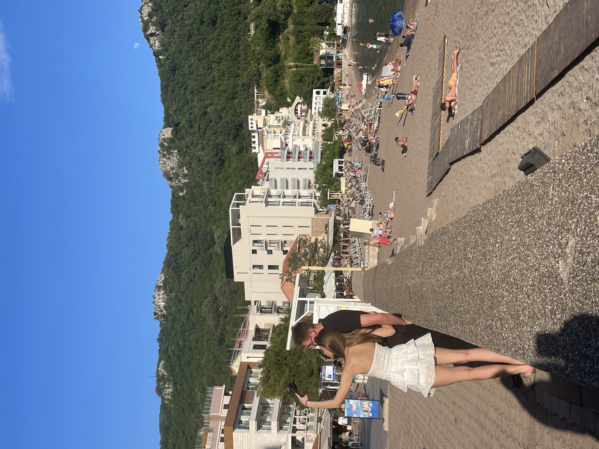 A medium-level, angled long shot captures a summer scene at Rafailovići, Montenegro, on a sunny day. The blue sky stretches from the upper left to the middle left, with a pale moon visible. A green, forested mountain occupies the left side of the frame. A series of white multi-story buildings line the middle-left, leading to a beachfront with closely packed beach chairs, umbrellas, and people. A wooden walkway is visible on the far right, with beachgoers scattered around.
A couple stands in the foreground. The woman, in a short white dress, extends her right arm holding a cell phone, seemingly taking a selfie. The man, dressed in a black shirt, wraps his arms around her waist. They stand on a paved walkway, with a low stone wall to the right. The couple are positioned towards the lower-middle section of the photo. The scene is bathed in strong sunlight, creating sharp shadows.