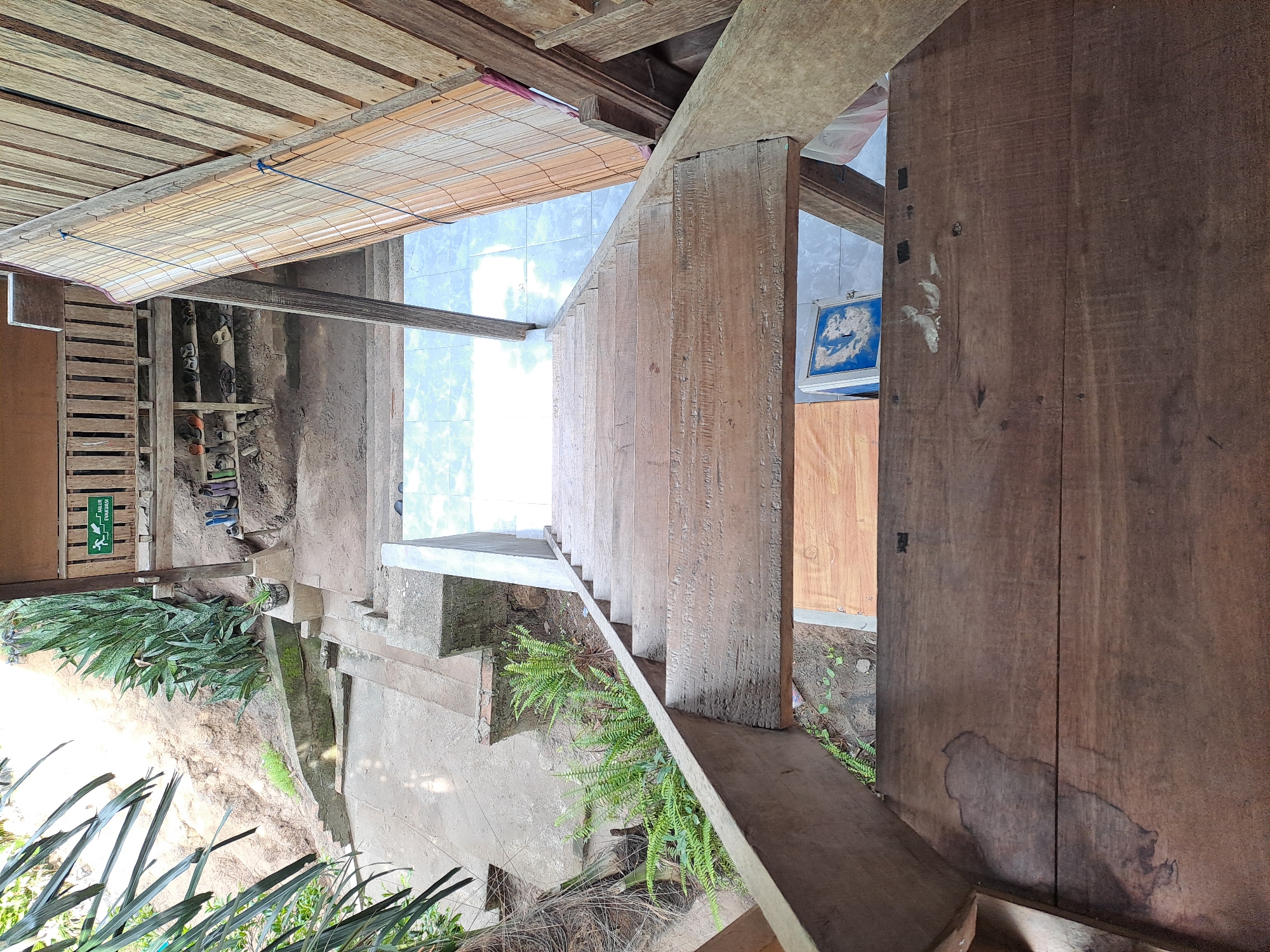 The image provides a top-down perspective of a wooden staircase leading down from a raised wooden structure. The staircase is made of weathered planks. Below the staircase, a tiled floor is visible. To the left of the staircase, a concrete pathway and a lush green fern are present, with a sandy embankment in the background. To the right, a bamboo blind hangs from the wooden structure. A green sign with white text and a pictogram indicating an evacuation route is mounted on a wooden railing of the structure, visible in the upper left portion of the image. Various pairs of shoes are arranged on shelves beneath the raised structure. The scene appears to be an outdoor or semi-outdoor area, possibly part of a dwelling or guesthouse. The location is identified as Geulanggang Baro, Indonesia.