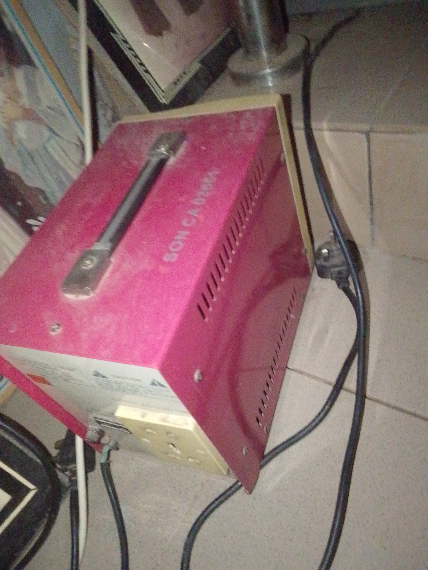 This is a medium-angle, slightly blurry, eye-level shot of a pink voltage stabilizer. The device is situated on a tiled floor, and to the left, partially visible, is a portion of a framed picture or artwork. The overall scene appears to be indoors, possibly a home or office setting, with the lighting suggesting daytime.
The voltage stabilizer is the primary subject. It has a metallic handle on top and vent slits along its side. There is visible dust on its surface. Text is present on the side of the stabilizer, appearing to read "SON CAM". On the front panel of the stabilizer, there are outlets and what seem to be warning labels. A black power cord is plugged into the side of the stabilizer and snakes away, disappearing out of frame. Another white cable is visible near the bottom left of the stabilizer.
The overall impression is one of a functional, albeit slightly dusty, piece of electrical equipment in use. The background is out of focus, with hints of other objects and a reflective metallic pole. There are no people visible in the image. The location context of Jalingo, Nigeria, is noted but not directly discernible from visual cues within the image itself.