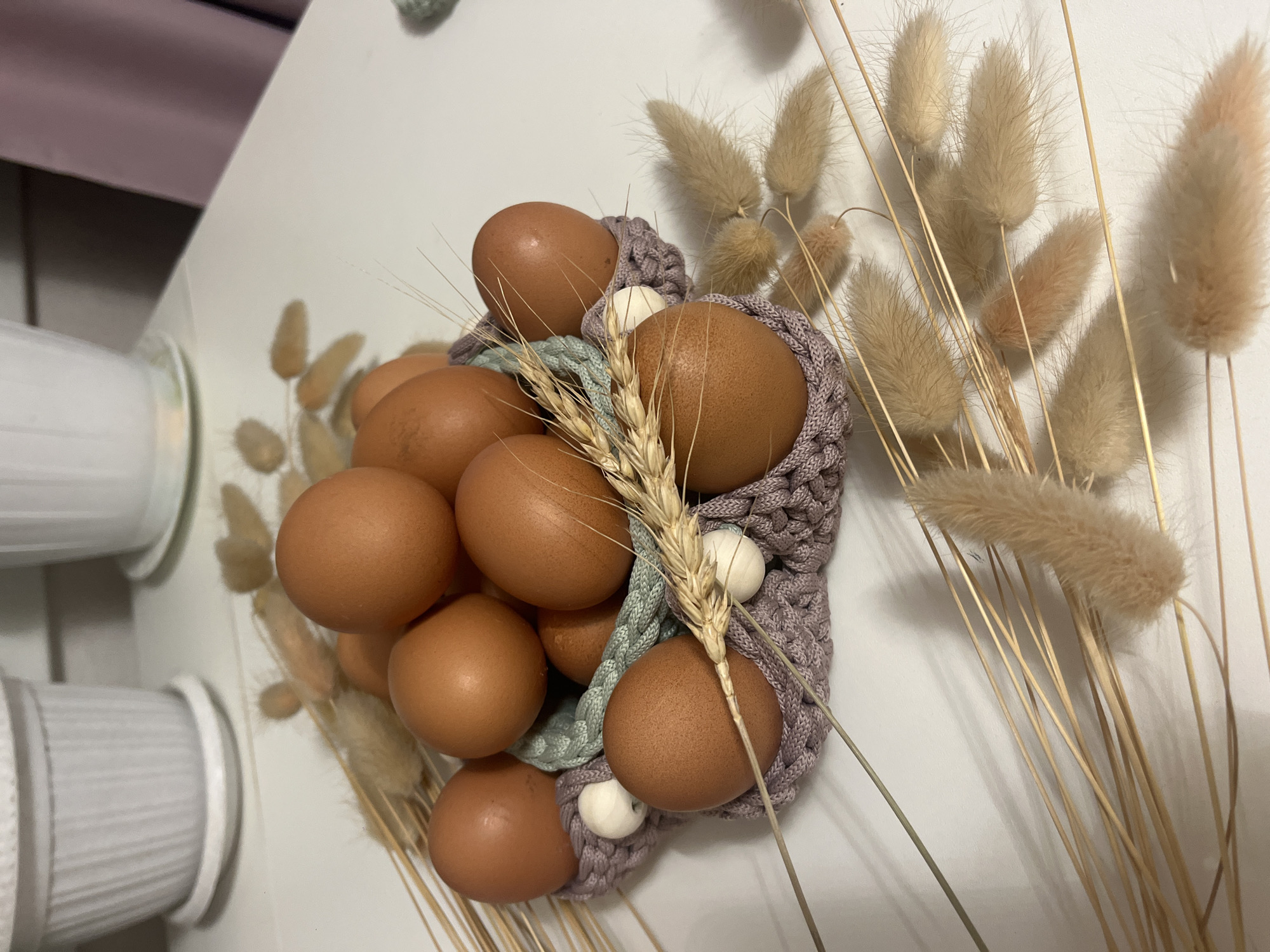 A still life photograph features a group of brown eggs nestled in a knitted or crocheted holder, adorned with sprigs of wheat and dried bunny tail grass. The holder is made of lavender and blue yarn. The arrangement is placed on a white surface.
The photograph does not appear to be associated with the location of Nizhny Novgorod, Russia.