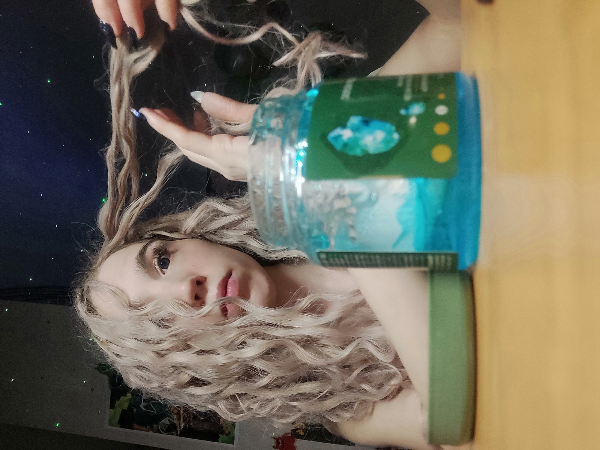 A woman with blonde curly hair is posing with a jar of blue hair gel. Her left hand is holding a strand of her hair. The jar has a green lid. The background behind her hair is dark with speckles of light. The jar is on a tan surface. The location is unknown, and there is no indication that the image was taken in Chaikovsky, Russia.