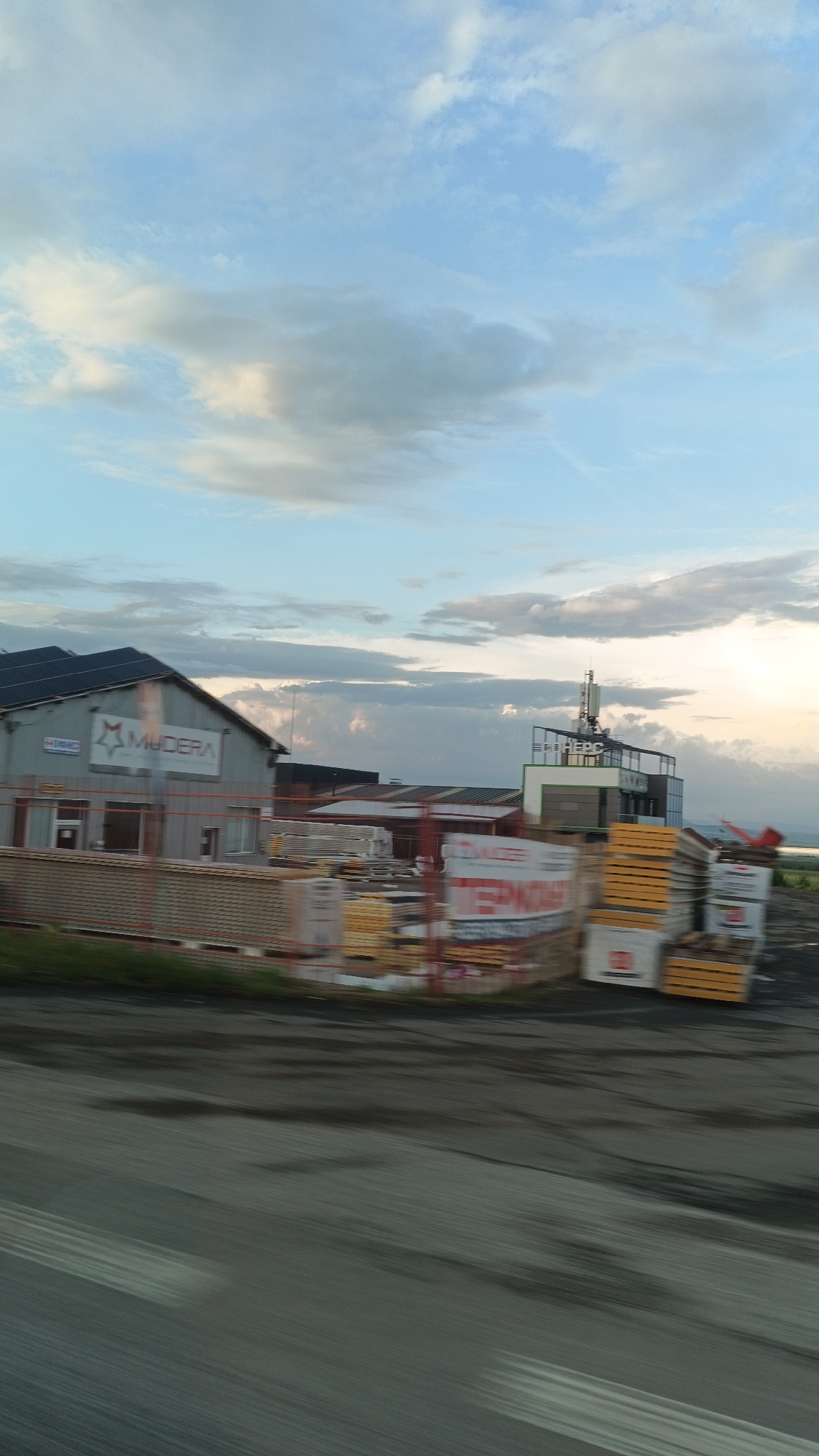 The image shows a street-level view in Burgas, Bulgaria, possibly taken from a moving vehicle. The foreground consists of a blurred asphalt road. To the left is a building with the sign "MUDERA" visible on its facade, with stacked lumber in front. Another building with a tower or antennae on top can be seen to the right of the "MUDERA" building, also with stacks of lumber in the foreground. The sky is partly cloudy with a mix of blue and grey hues. The scene gives the impression of an industrial or commercial area with lumber processing or sales facilities. The general setting is slightly hazy, possibly indicating dusk or dawn.