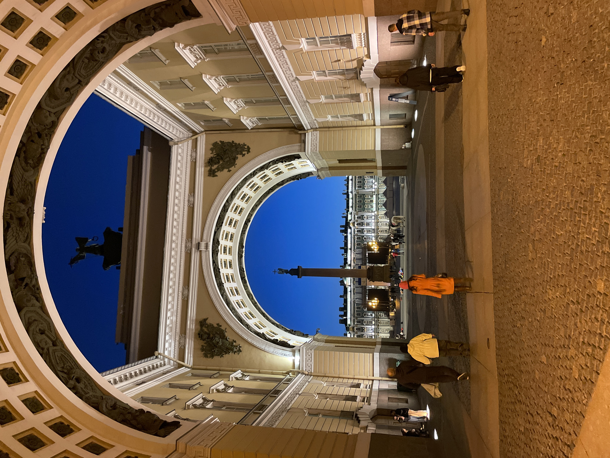 The image presents a grand architectural scene at dusk or night, viewed through a massive, ornate archway. The sky above is a deep, clear blue, contrasting with the warm, artificial light illuminating the surrounding buildings and arch. The archway itself is a dominant feature, constructed of light-colored (beige/cream) stone with intricate white classical details. Its inner curve is richly decorated with detailed carvings, including what appear to be winged mythological figures and decorative motifs.
Through this monumental arch, a large, open square or plaza stretches into the distance. A tall, slender column, reminiscent of the Alexander Column, stands prominently in the middle distance, topped with a statue. Beyond the column, another large, illuminated building, likely a palace or significant historical structure, completes the background.
Several individuals are present, providing a sense of scale. In the immediate foreground on the right, a person in a plaid shirt and dark pants stands beside another in a dark jacket. Further into the square, a person wearing a bright orange jacket and a red hat is visible, along with others in various jackets, walking or observing the scene. The ground is paved, with textured cobblestones or similar blocks on the right foreground, transitioning to smoother flagstones nearer the arch. The overall impression is one of historical grandeur and architectural beauty, captured during the twilight hours. No visible text is discernible in the image.