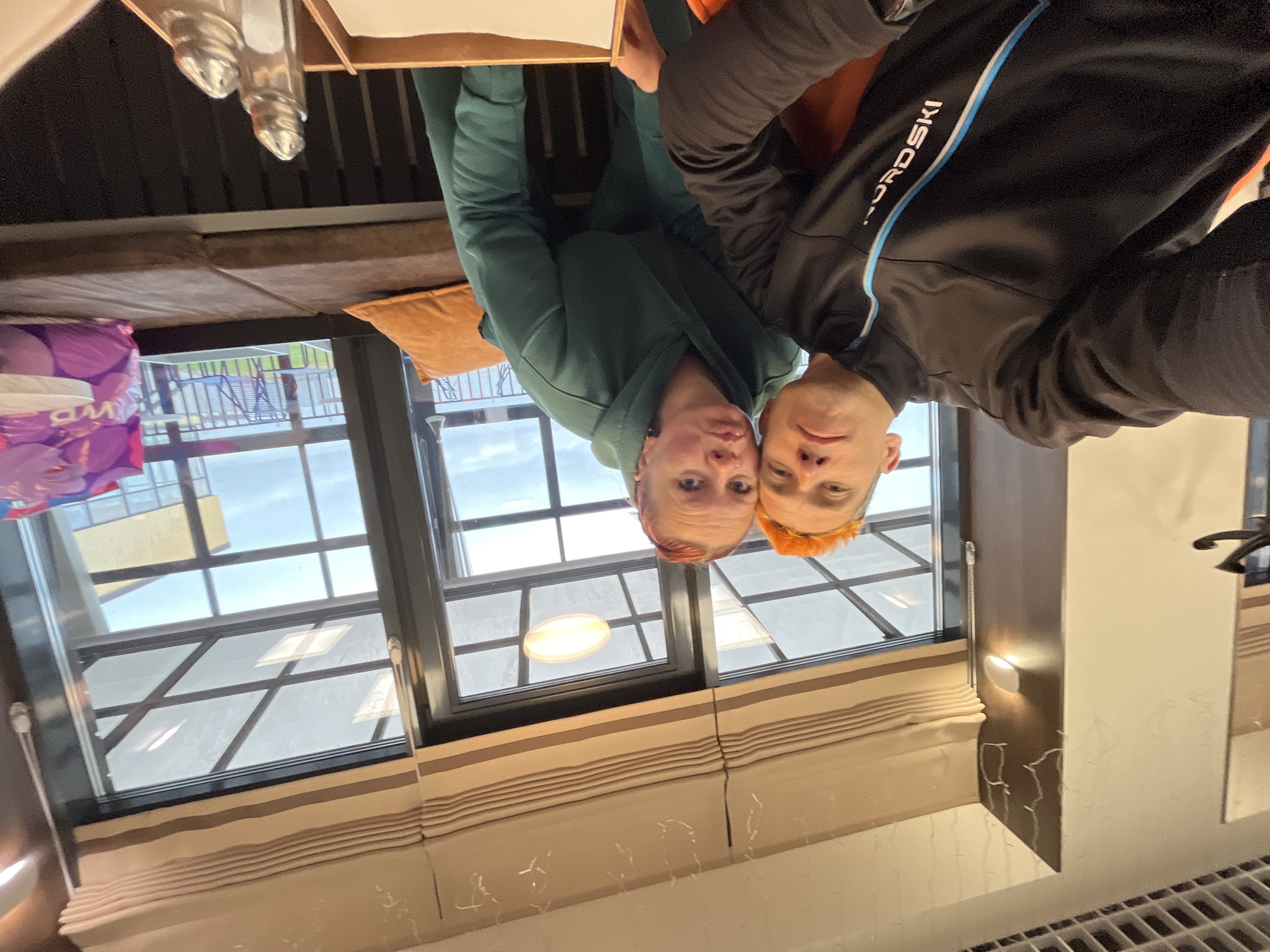 Two individuals are visible in an indoor setting. One is wearing a green hoodie, the other is in a black jacket with a white and blue stripe and the word "NORDSKI" on the sleeve. The background features a window with a view of a building and a sky with clouds. Inside the room, there is a booth with a brown cushion, a pillow, a table, and possibly salt and pepper shakers. There is also a purple bag with white text. The location might be in the city of Chaikovsky, Russia, based on context, but this cannot be determined from the image alone.