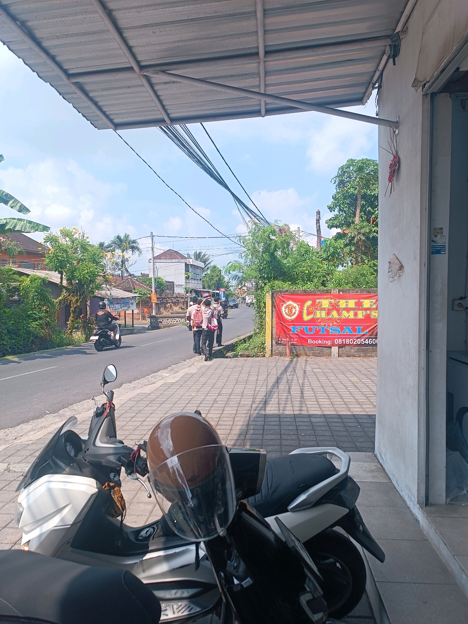 A street-level view captures a sunny outdoor scene in Tabanan, Indonesia. In the foreground, two scooters are parked on a tiled sidewalk. The scooter on the left is partially visible, with its front wheel and handlebars showing, a brown helmet resting on the seat. The scooter on the right is more fully visible, black and white, with its seat and rear fender. Beyond the sidewalk, a paved road stretches into the distance, with a white line marking the center.
Several people are present on the road. A person on a white scooter rides away from the camera, wearing a dark shirt and helmet. Further down the road, a group of four young people, appearing to be students in uniform with backpacks, walk along the side of the road. A red banner with text in yellow and white is visible on the right side of the road. The banner reads "THE CHAMP'S FUTSAL" and includes a phone number for bookings. Buildings and lush green trees line the sides of the road, with houses and more distant structures also visible. Overhead, electrical wires are strung across the frame, supported by poles, and a corrugated metal roof extends from a building into the foreground. The sky is blue with scattered white clouds.
