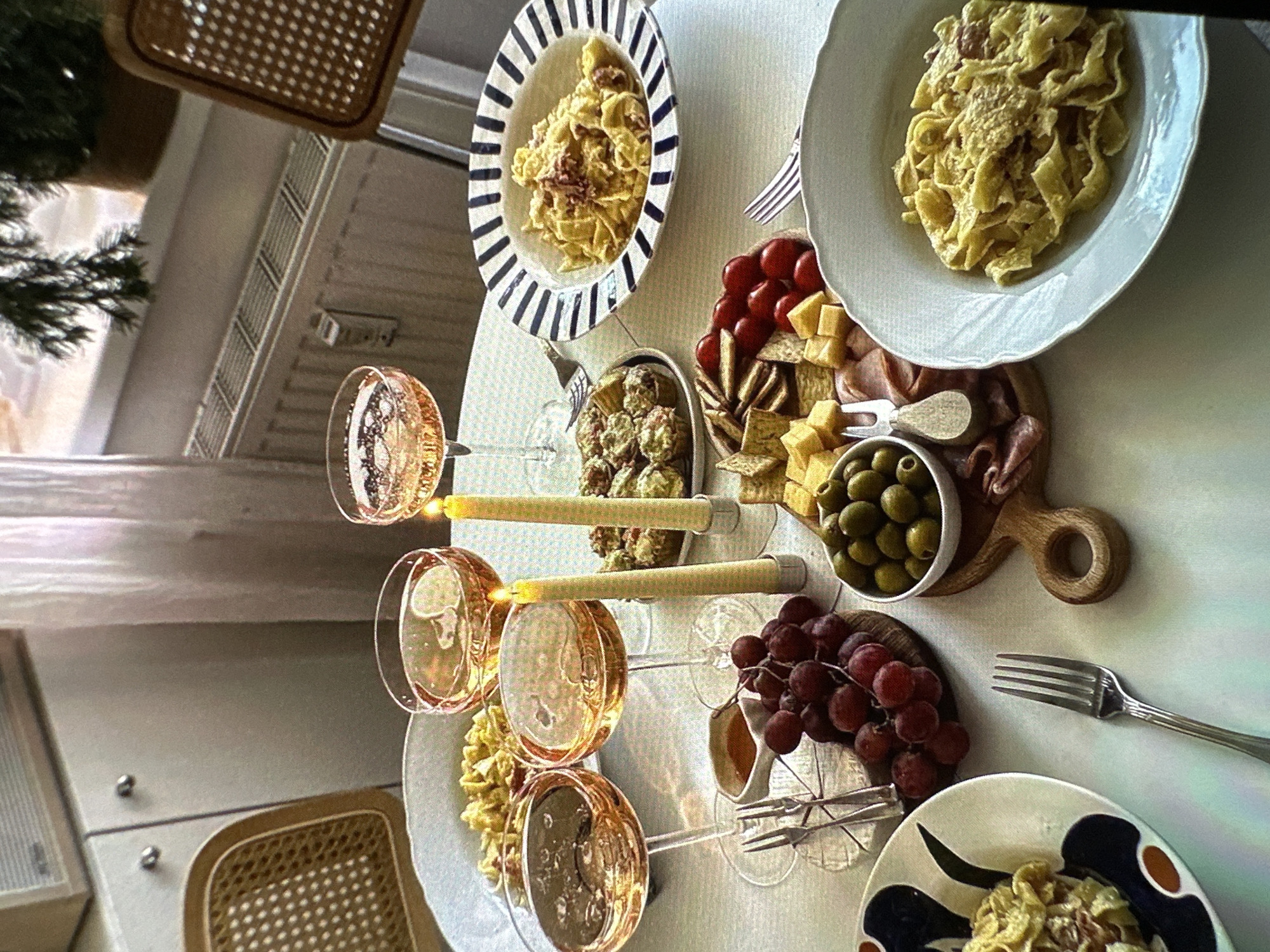 A table is set for a meal with plates of pasta, a charcuterie board, grapes, olives, and other items. There are three wine glasses with a candle between each glass. The scene appears to be indoors, possibly in a dining room or kitchen. The location is not determinable. No people are visible in the image. The general atmosphere suggests a planned or staged setting.