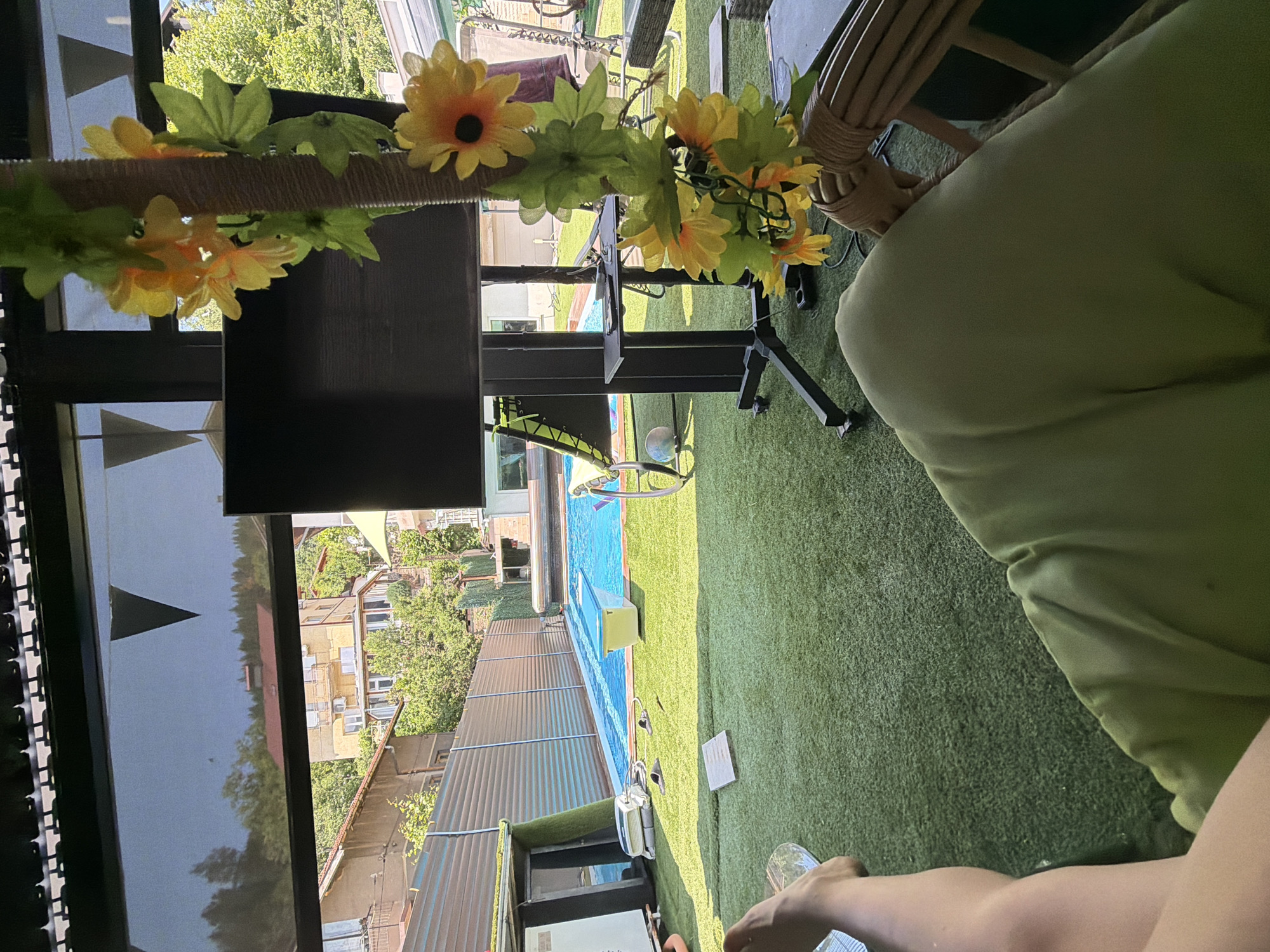 The image shows a person's legs and lap covered by a light green fabric, in the foreground. The scene is an outdoor patio with artificial turf covering the ground. In the background, there is a swimming pool with bright blue water and a yellow rectangular object floating in it. Beyond the pool, a metal fence is visible, and further back, buildings and trees are seen, suggesting an urban or suburban setting. A large black television screen on a stand is also present, decorated with artificial sunflowers and green leaves. The overall ambiance suggests a leisure setting, possibly a backyard or a poolside area. The image does not contain enough information to determine the specific location in Sofia, Bulgaria.