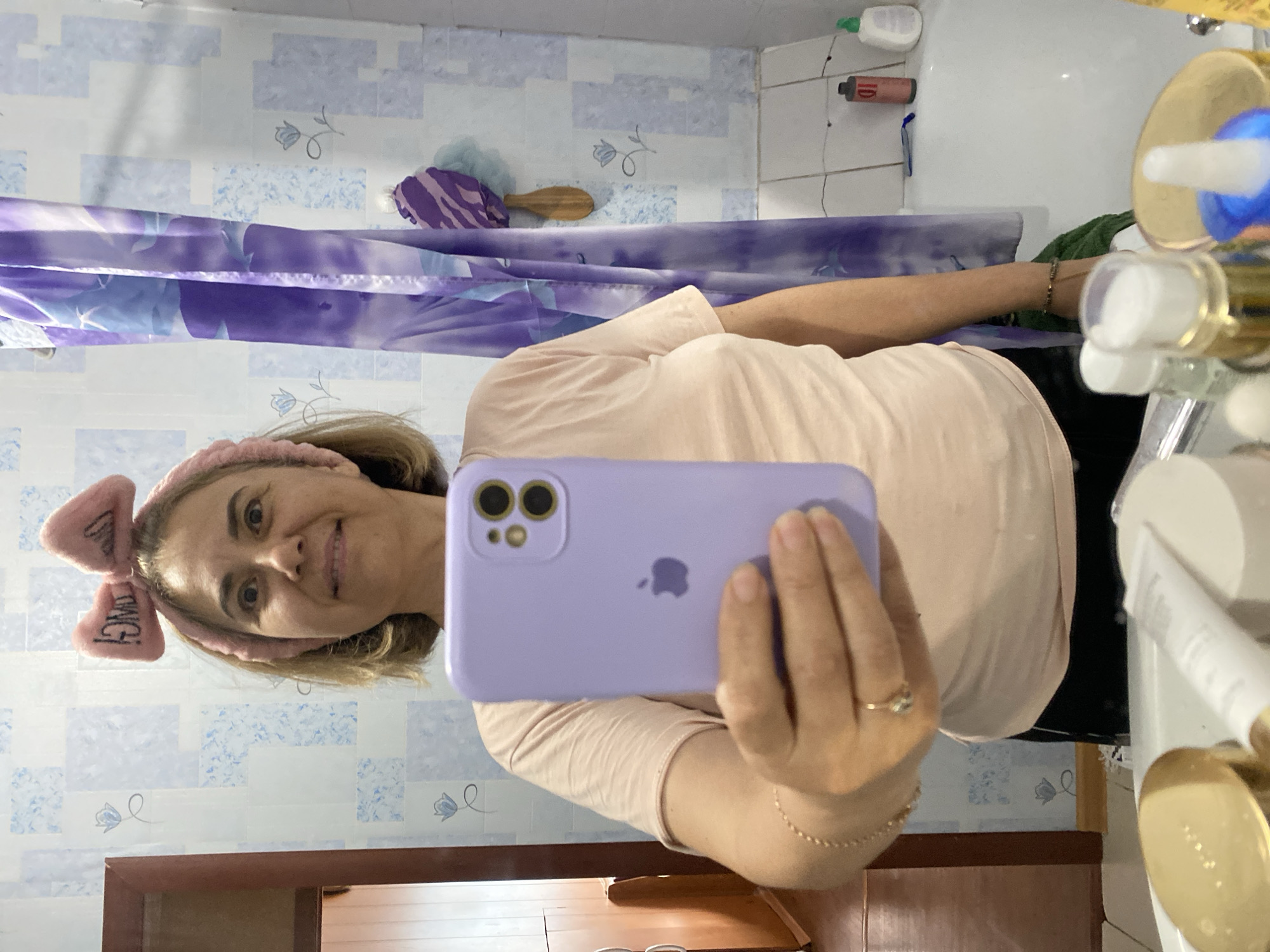 A woman with light brown hair tied back with a pink headband with a bow on top is taking a mirror selfie in a bathroom. She is wearing a light pink t-shirt and black bottoms. She is holding a purple iPhone in her right hand. The bathroom has blue and white patterned tiles on the wall and a purple shower curtain. There are various toiletries visible on the counter in the foreground, including bottles and jars. The setting appears to be a private residence in the city of Urazovo, Russia.