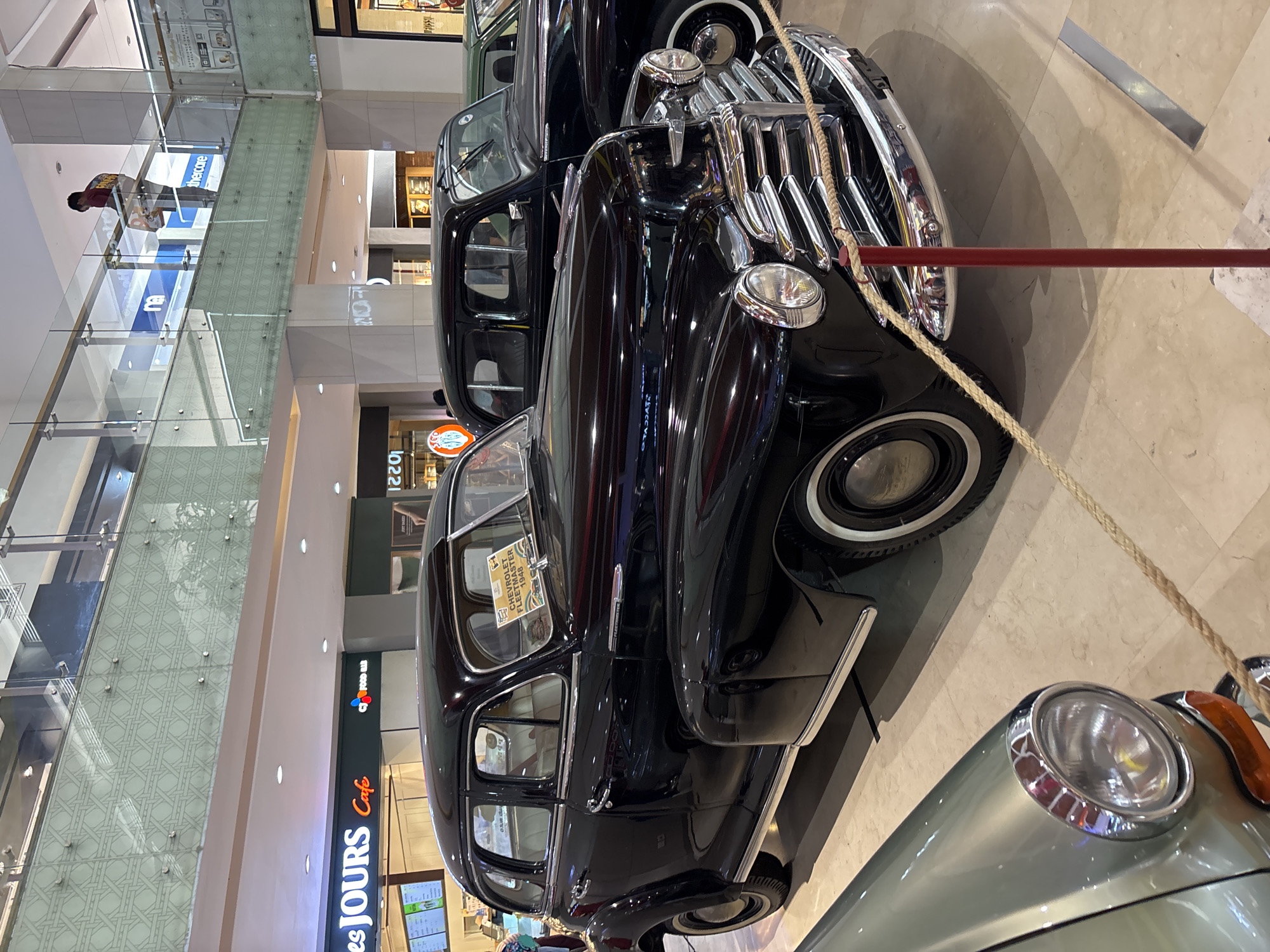 The image shows two vintage cars displayed in an indoor mall. The car closest to the viewer is a black four-door sedan with chrome detailing and whitewall tires. A sign is visible on the windshield, indicating it is a "Chevrolet Fleetmaster 1948." A rope is draped across the front of the car, presumably to prevent access. In the foreground, the headlight of another vehicle is partially visible.
The mall environment is discernible, featuring glass railings, multiple floors, and retail signage. A restaurant named "es JOURS Cafe" is partially visible. The reflection in the glass suggests the location could be in Kuta, Indonesia, but there is no definitive proof. People are visible on upper floors of the mall.