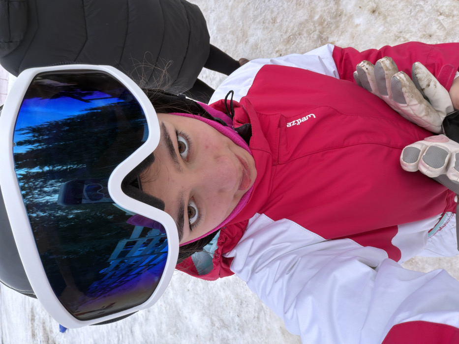 An outdoor scene in Belmont, France, depicts a young female individual from the chest up, positioned centrally and looking directly at the camera with her tongue slightly extended. She wears a black ski helmet and white-framed ski goggles with highly reflective blue lenses; these lenses reflect tall coniferous trees, a multi-story building with multiple windows, and a vertical snowboard. A pink balaclava or neck warmer is visible around her lower face and neck. She is dressed in a red and white ski jacket, with the brand "wed'ze" visible on the right chest. In her left hand, she holds a pair of light-colored, soiled ski gloves with grey textured palms. The ground surrounding her is covered in patchy, dirty snow. A second individual, wearing a black puffer jacket and dark pants, is partially visible standing in the upper right background.