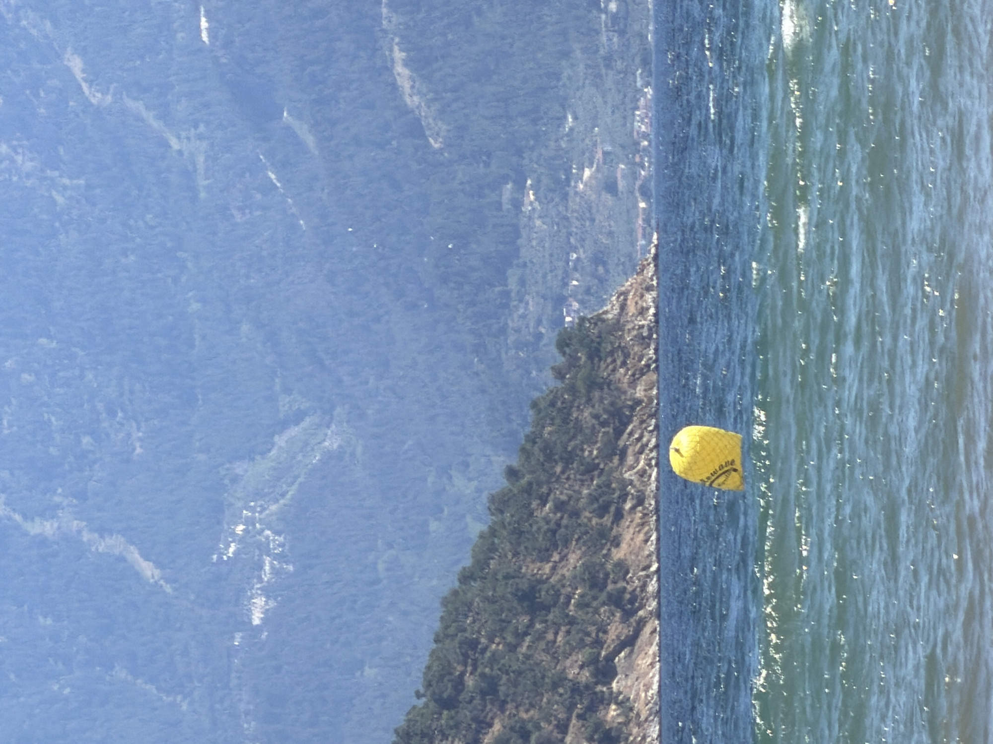 A yellow buoy floats on the surface of choppy blue water. The buoy is cylindrical with a rounded top and has some text visible on its side. In the background, a steep, tree-covered hillside slopes down towards the water, with a rocky shoreline. Further in the distance, the hazy outline of a mountain range covered in dense forest is visible against a pale blue sky. The scene depicts a coastal area, likely in or near Keramoti, Greece, given the characteristic Mediterranean landscape. There are no people or animals present in the image.