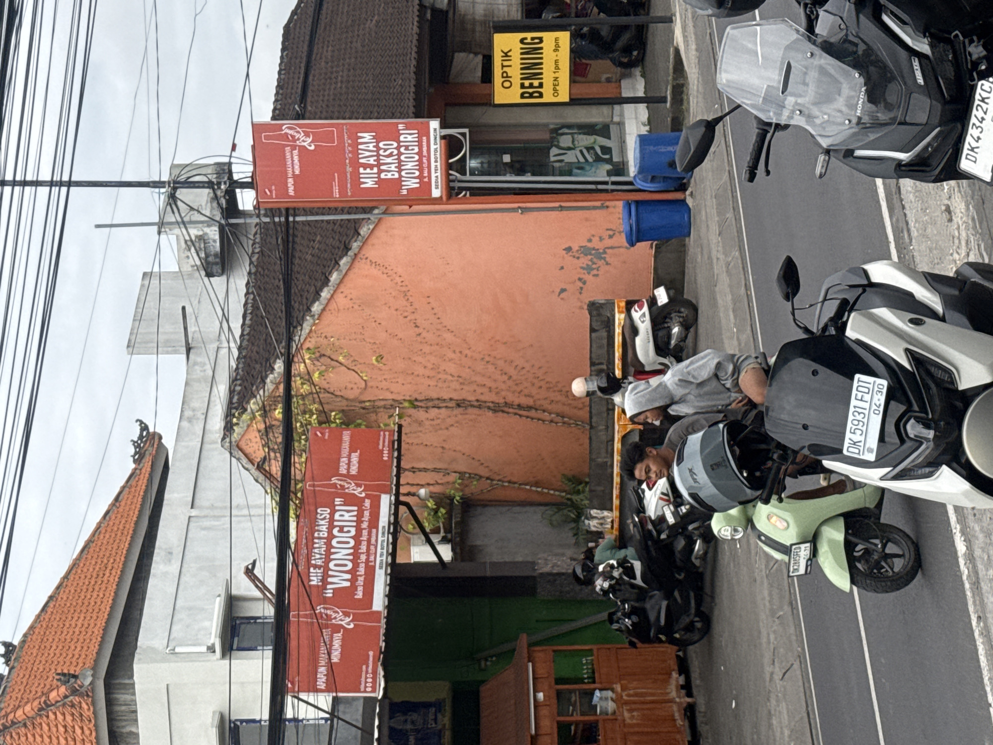This is a vertical, eye-level, medium shot of a street scene in Kuta Selatan, Indonesia. In the foreground, several parked motorcycles are visible, including a white and black scooter with the license plate DK 5931 FDT and a lighter green scooter with the license plate DK 2885 FED. One person, wearing a gray hoodie, sits on one of the motorcycles. In the background, there is an orange building with a patterned vine growing on it. Two red signs with white text are affixed to the building. The upper sign reads "MIE AYAM BAKSO 'WONOGIRI'" and the lower sign repeats this text with additional details. A yellow sign with black text above the orange building reads "OPTIK BENNING OPEN 1pm-9pm". The sky is overcast, suggesting it might be daytime. Electrical wires crisscross the upper portion of the image. Two blue buckets are visible on the street.