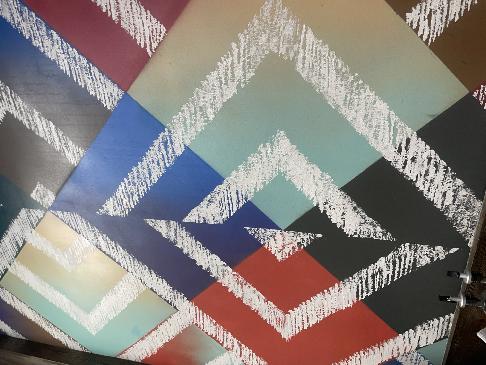 The image is a close-up, abstract shot of a wall with a geometric pattern. The pattern consists of intersecting diagonal lines and rhombuses in various muted colors such as teal, yellow, blue, red, gray, and brown. White, textured brush strokes overlay these colored sections, creating a zigzag or chevron-like effect within the rhombuses. The lighting is diffuse, and the focus is sharp on the textured surface of the wall. The bottom of the image shows the top of two silver pour spouts attached to glass bottles, suggesting the wall is part of a bar or restaurant setting. The scene is devoid of people or any discernible landmarks that would indicate a specific location in Tashkent, Uzbekistan.