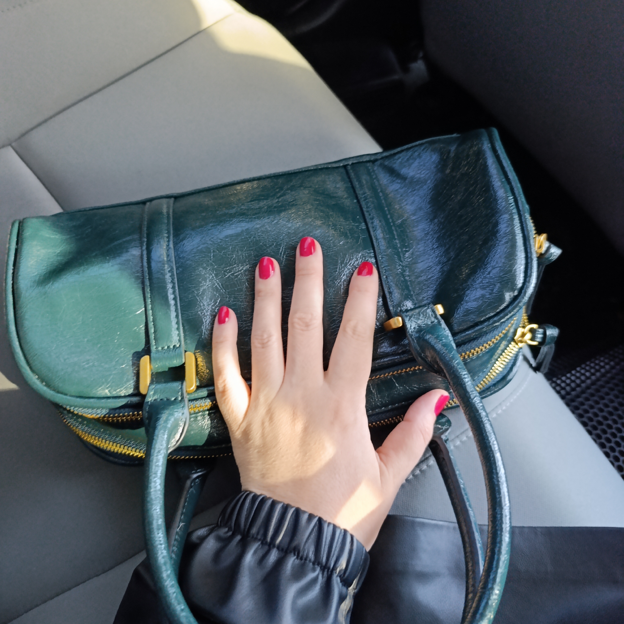 This image, taken in the city of Марковский, Russia, shows a close-up, high-angle view of a human hand resting on a dark green handbag inside a vehicle. The hand, presumed female, has light-toned skin and all visible fingernails are painted bright red. It is positioned centrally, palm down, with fingers slightly splayed over the bag's surface. The handbag is a structured, rectangular item with a glossy, possibly patent leather-like finish, featuring gold-toned zippers and hardware, including visible buckles and rings. A dark, possibly black, sleeve with an elasticated cuff is visible around the wrist of the hand, indicating the individual's attire. The background consists of a vehicle interior, specifically a two-toned car seat upholstered in light grey and dark grey fabric. Natural light enters from the left, casting a shadow of the hand onto the handbag.