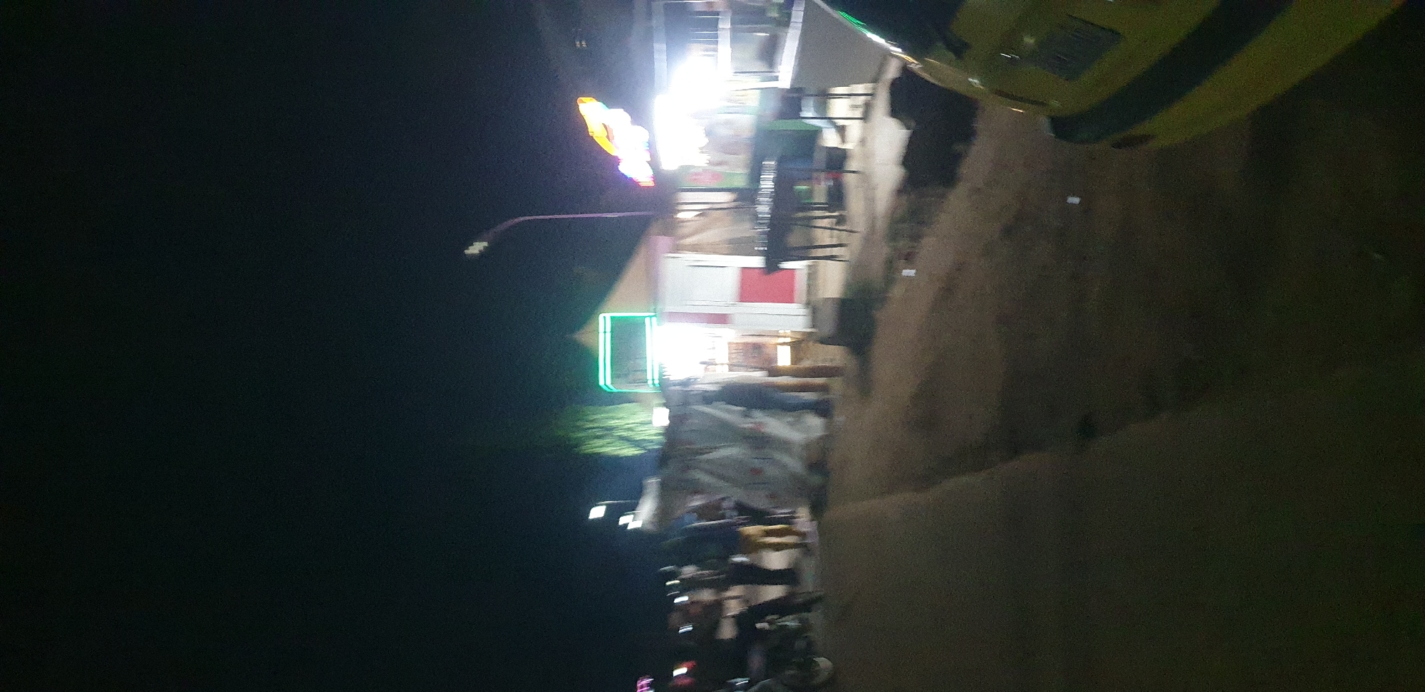 This is a nighttime street scene in Narayi, Nigeria, showing a commercial area with several structures and people. In the foreground, the rear of a yellow vehicle is visible on the right side. The middle ground features a collection of makeshift stalls and businesses. One stall has a bright green illuminated sign and red and white paneling. Another stall displays a bright, possibly orange, neon sign. Various individuals are present, some standing near the stalls, while others appear to be walking or engaged in activities. In the background, a street lamp is visible. The ground is unpaved, with a dirt or gravel surface. The overall atmosphere suggests a bustling night market or roadside commercial activity.